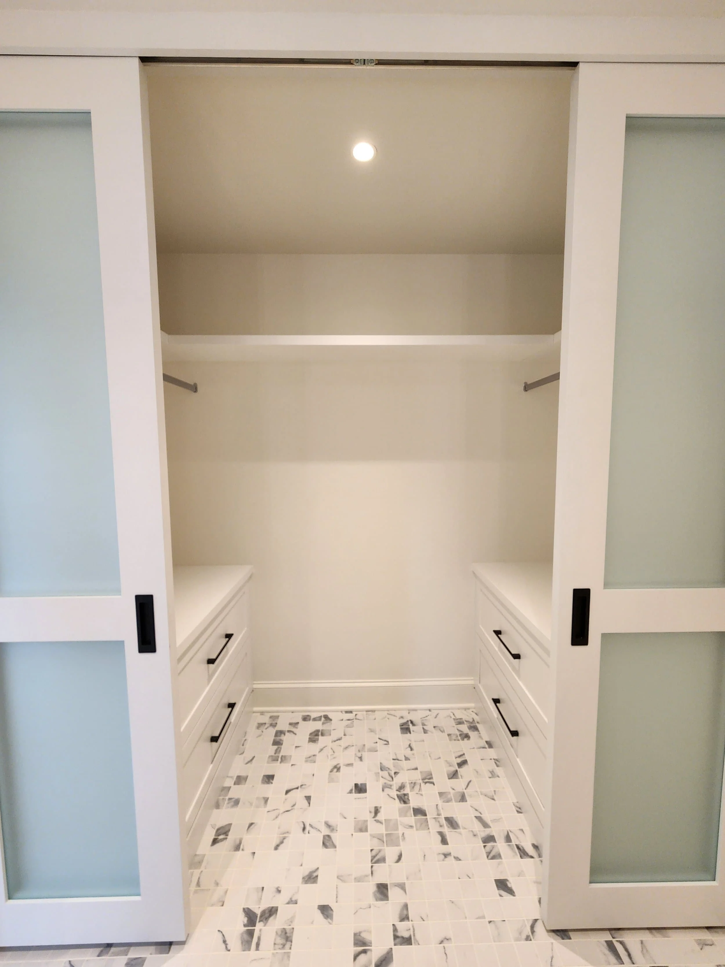Empty walk-in closet with white framing, sliding doors with frosted glass, interior shelving, and drawers, with marble-patterned tile flooring and a ceiling light.