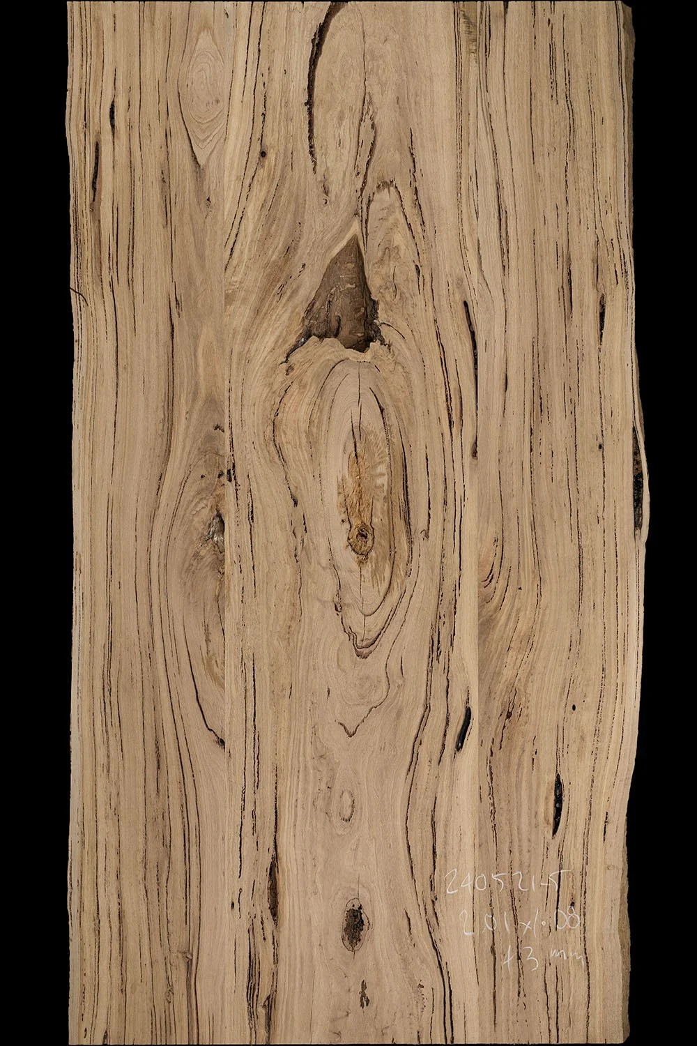 A close-up of a wooden slab with natural grain patterns and knots, some handwritten measurements and notes in white ink on the bottom right corner.