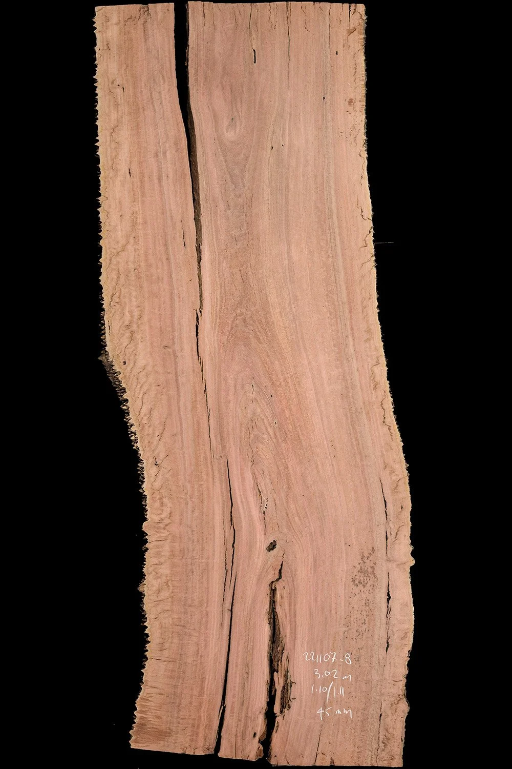 A slice of light-colored wood with natural grain patterns and some dark cracks, placed against a black background. Handwritten notes are visible on the bottom right side.