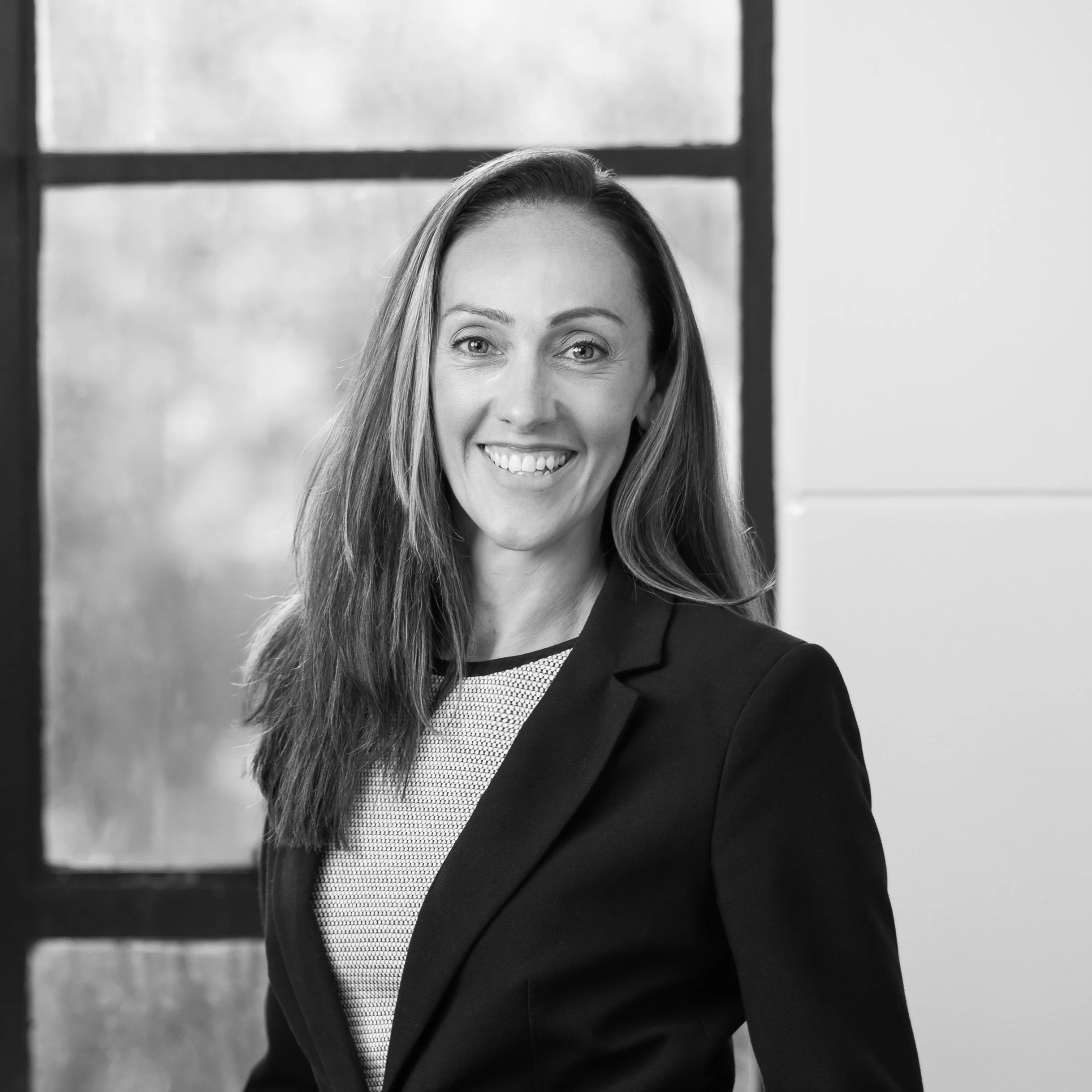 Black and white portrait of a woman with long hair, wearing a blazer, smiling, in an indoor setting with large window in the background.