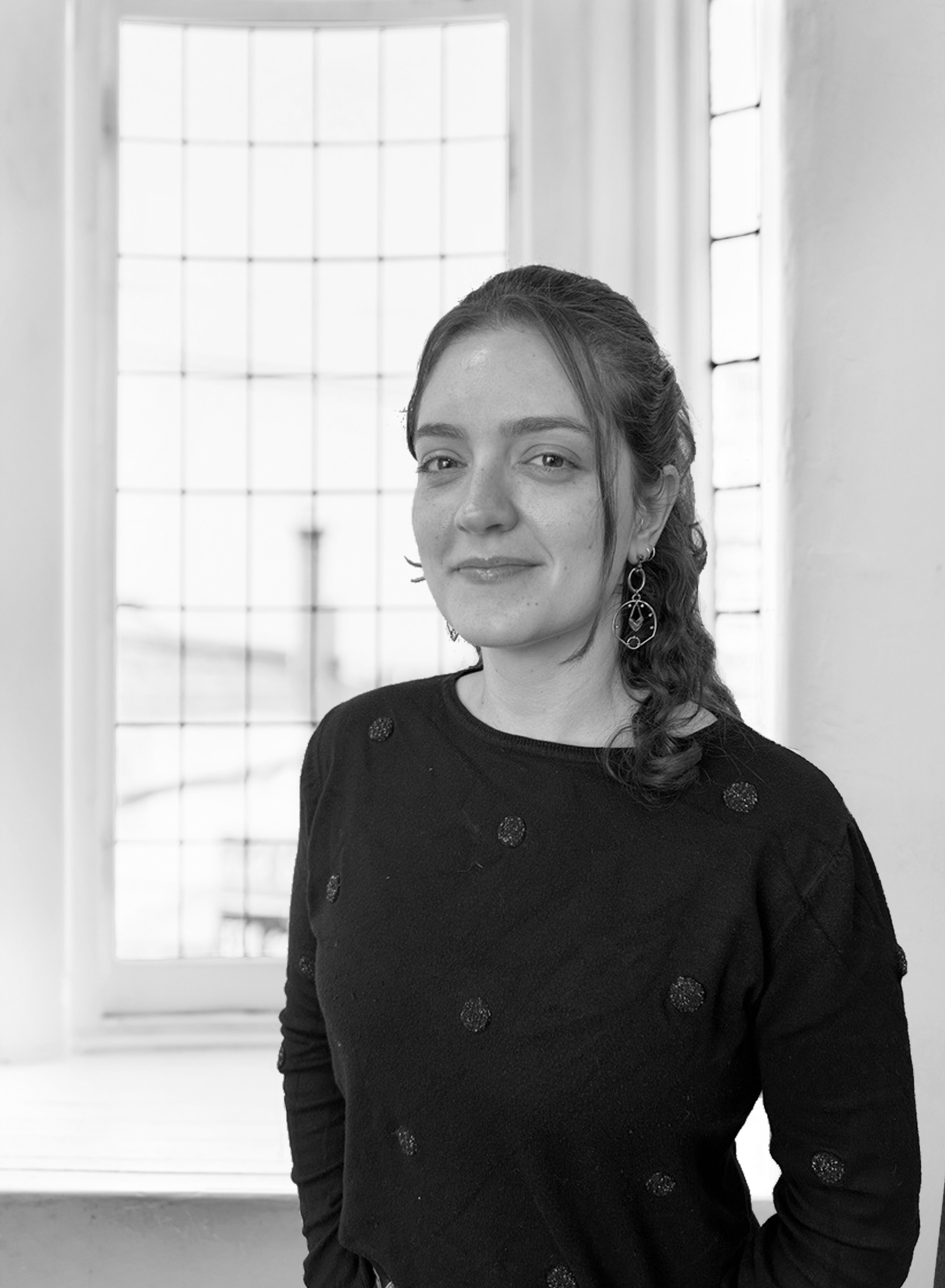 Portrait of a woman with dark eyes and curly hair, wearing a black sweater with decorative dots, standing indoors near large window with grid pattern, in black and white.