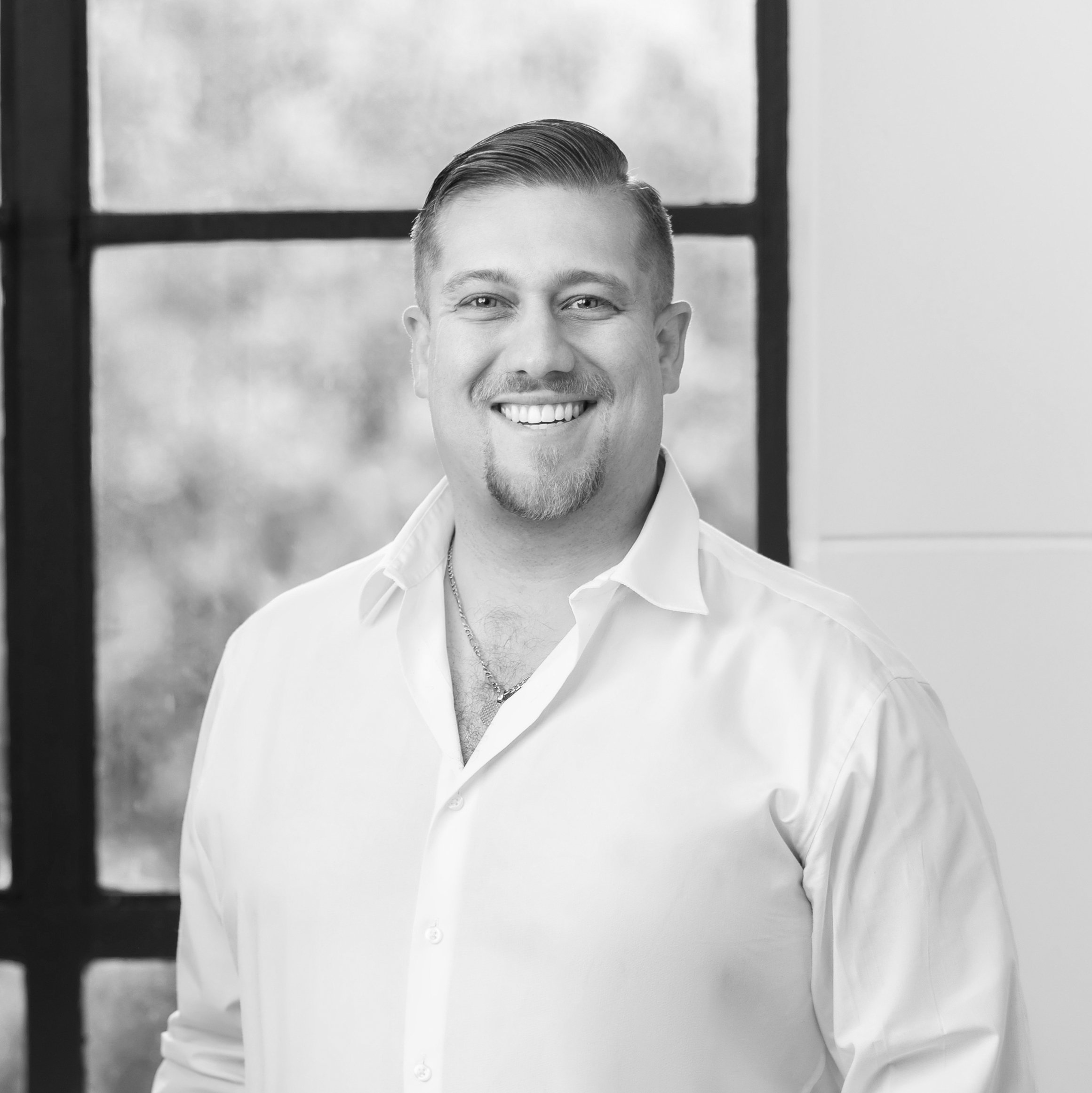 Black and white portrait of a smiling man with short, slicked-back hair wearing a white button-up shirt, standing in front of a window with trees outside.