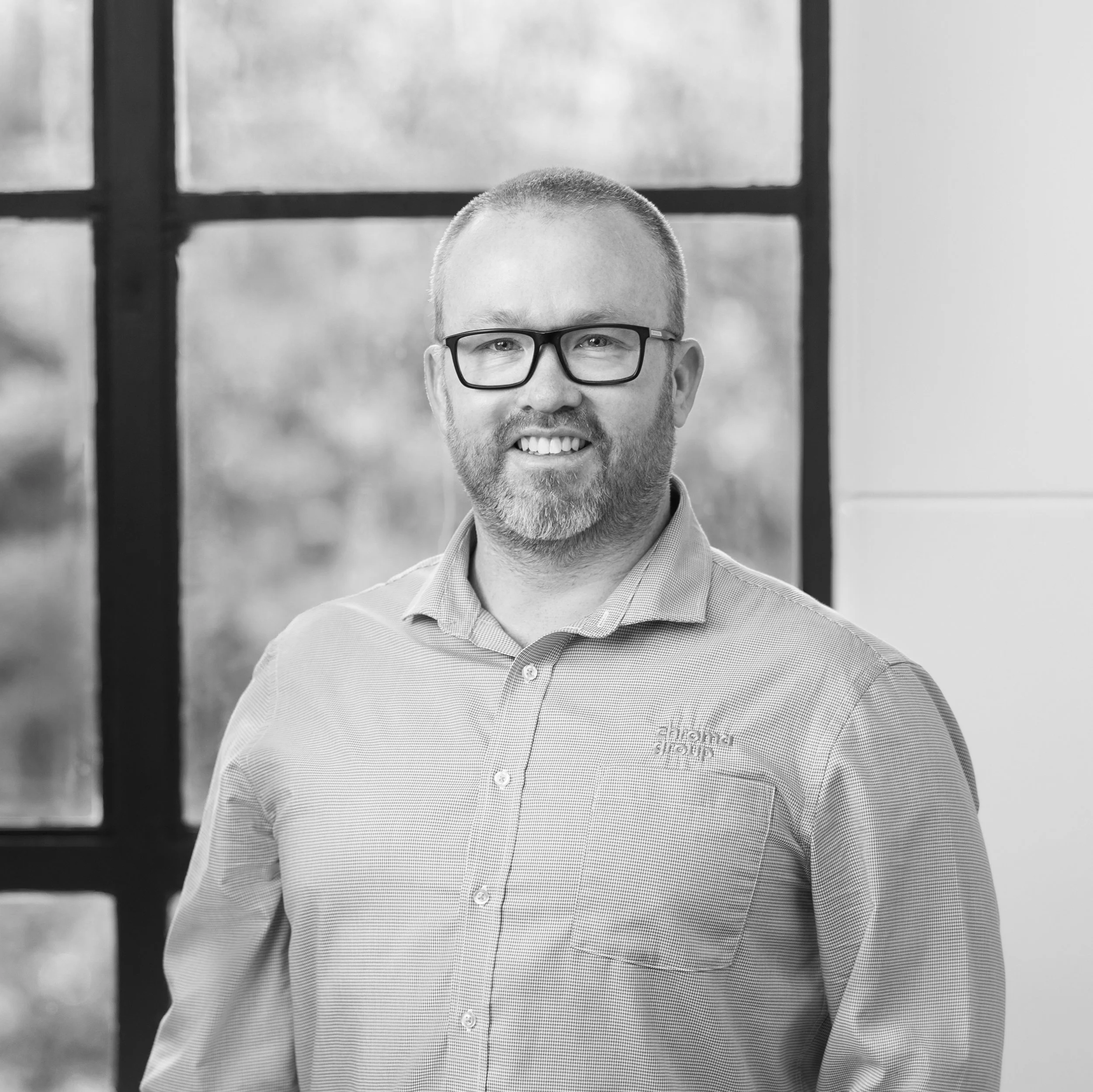 Portrait of a man with glasses, a beard, and short hair, smiling while standing indoors near a window.