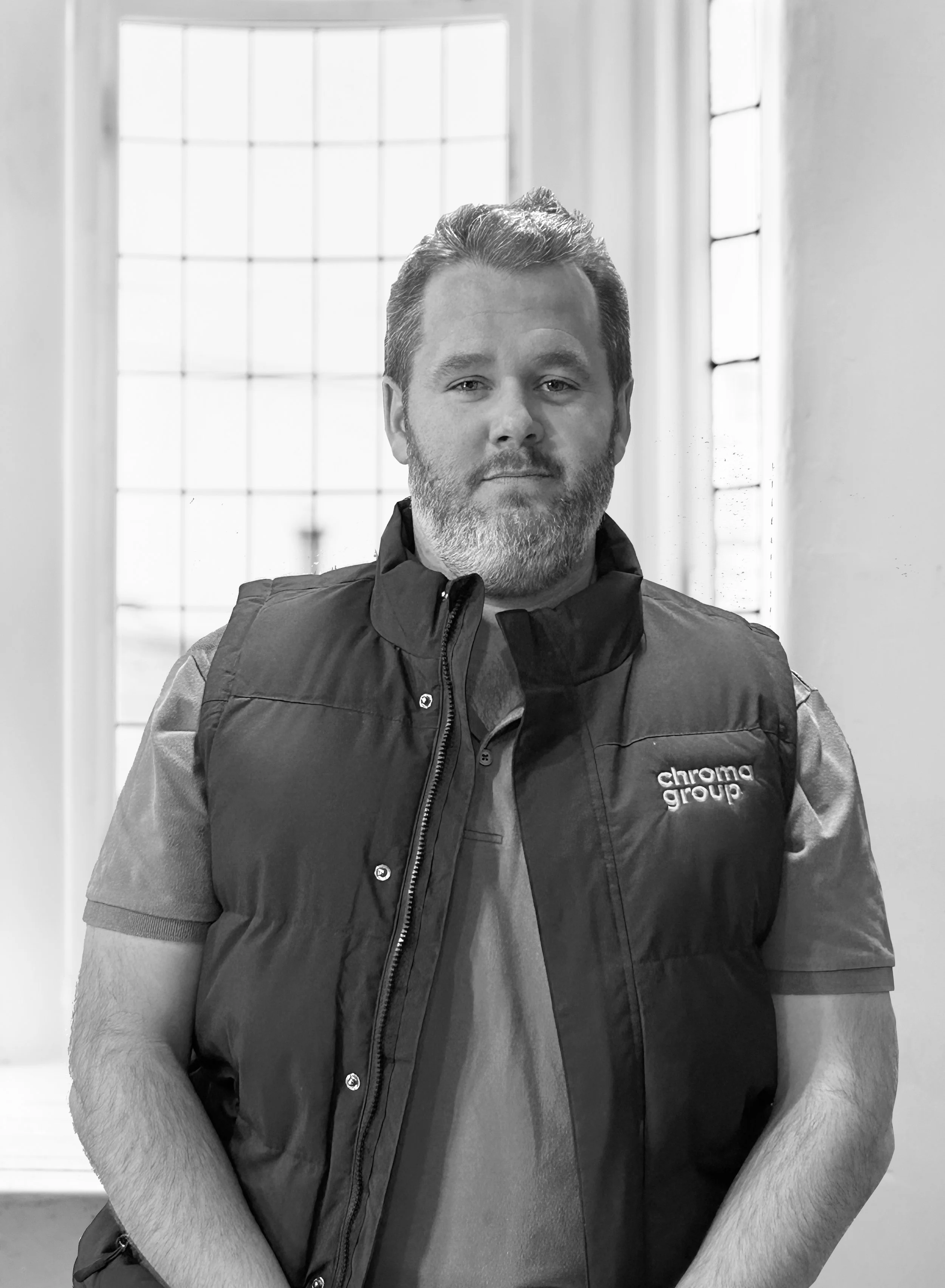 A man with a beard wearing a shirt and a vest with the logo 'chroma group' stands indoors against a windowed wall.
