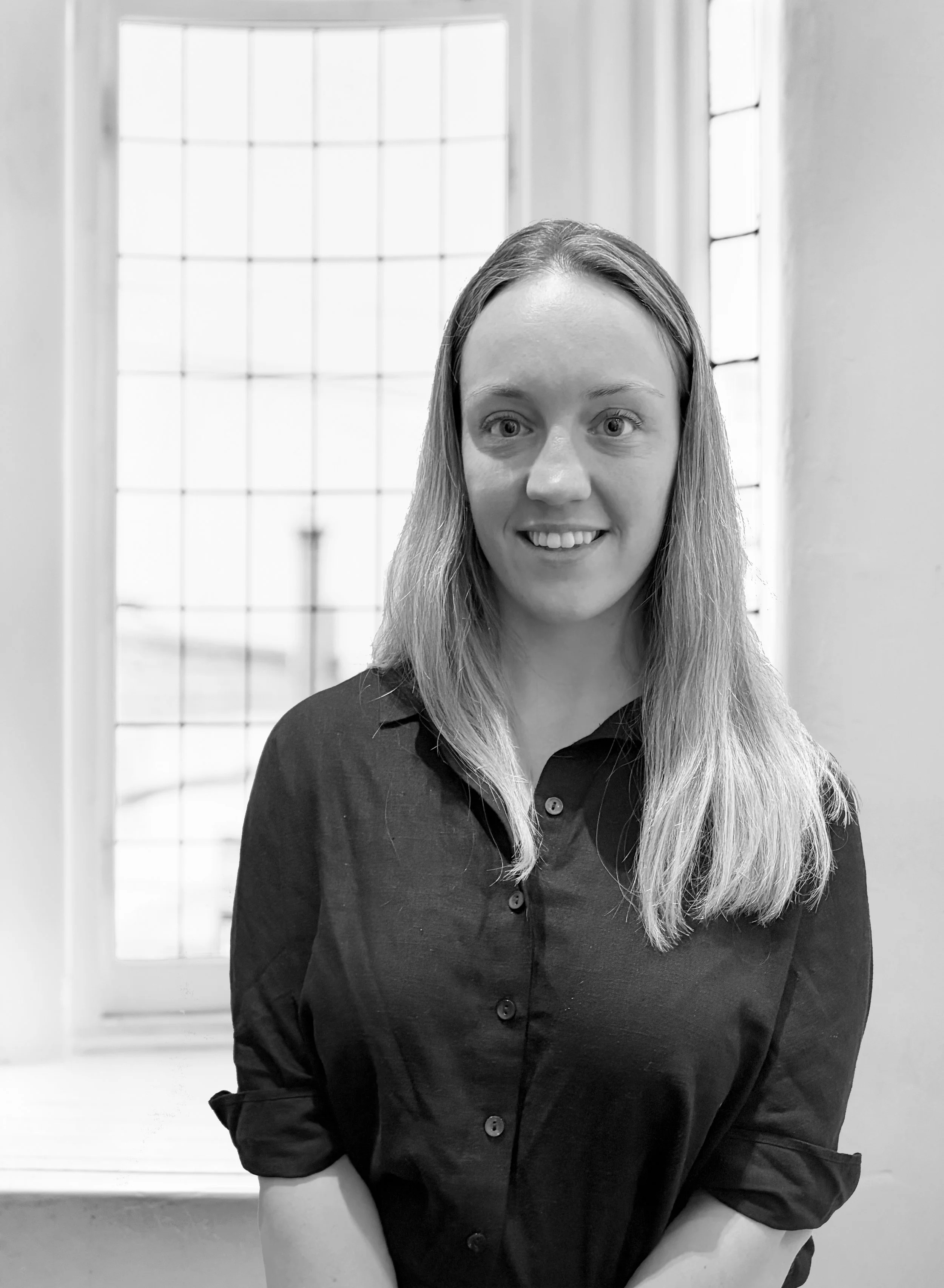 Black and white photo of a woman with long, straight hair, wearing a dark button-up shirt, standing in front of a window with grid pattern.