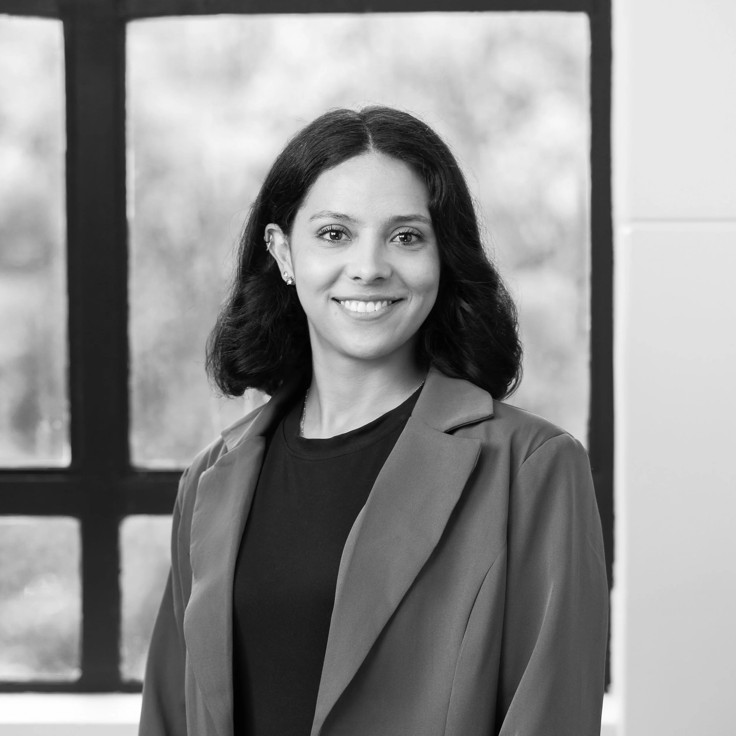 A black-and-white portrait of a smiling woman with shoulder-length dark hair, wearing a blazer and a dark top, standing indoors near large windows.