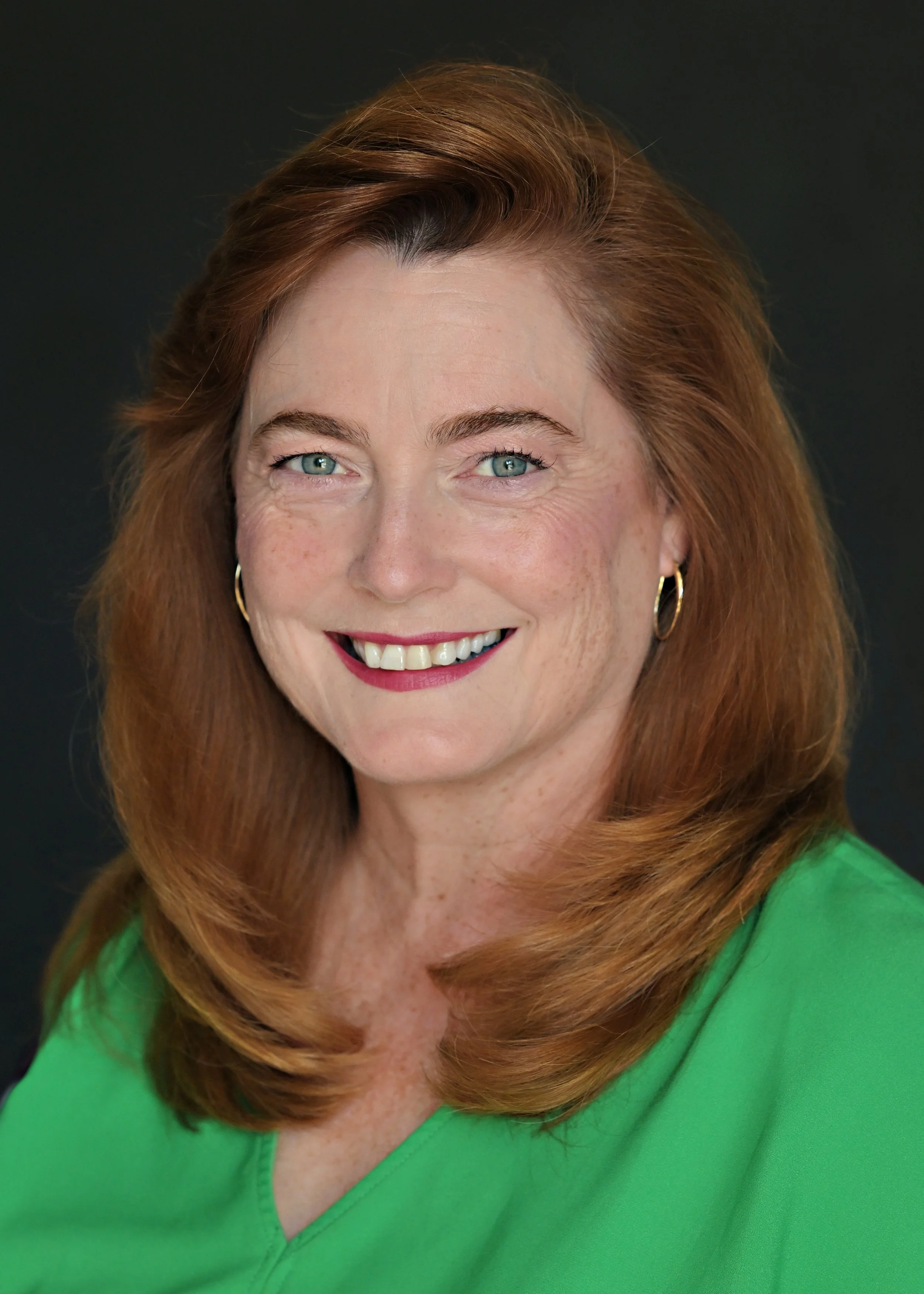 Close-up portrait of a woman with reddish-brown hair, wearing a dark gray sleeveless top, standing indoors with a white paneled wall in the background.