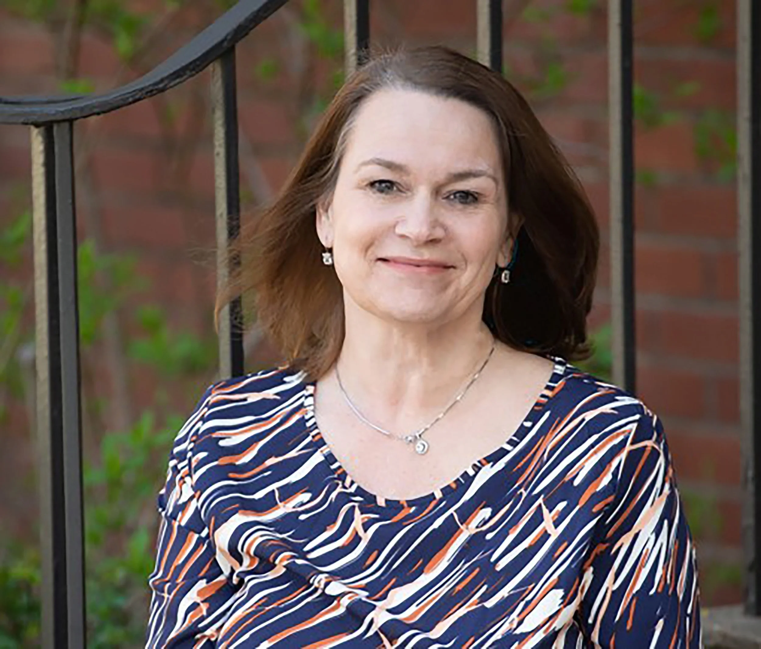 A woman with brown hair wearing a black blazer, necklace, and earrings, against a blurred background.