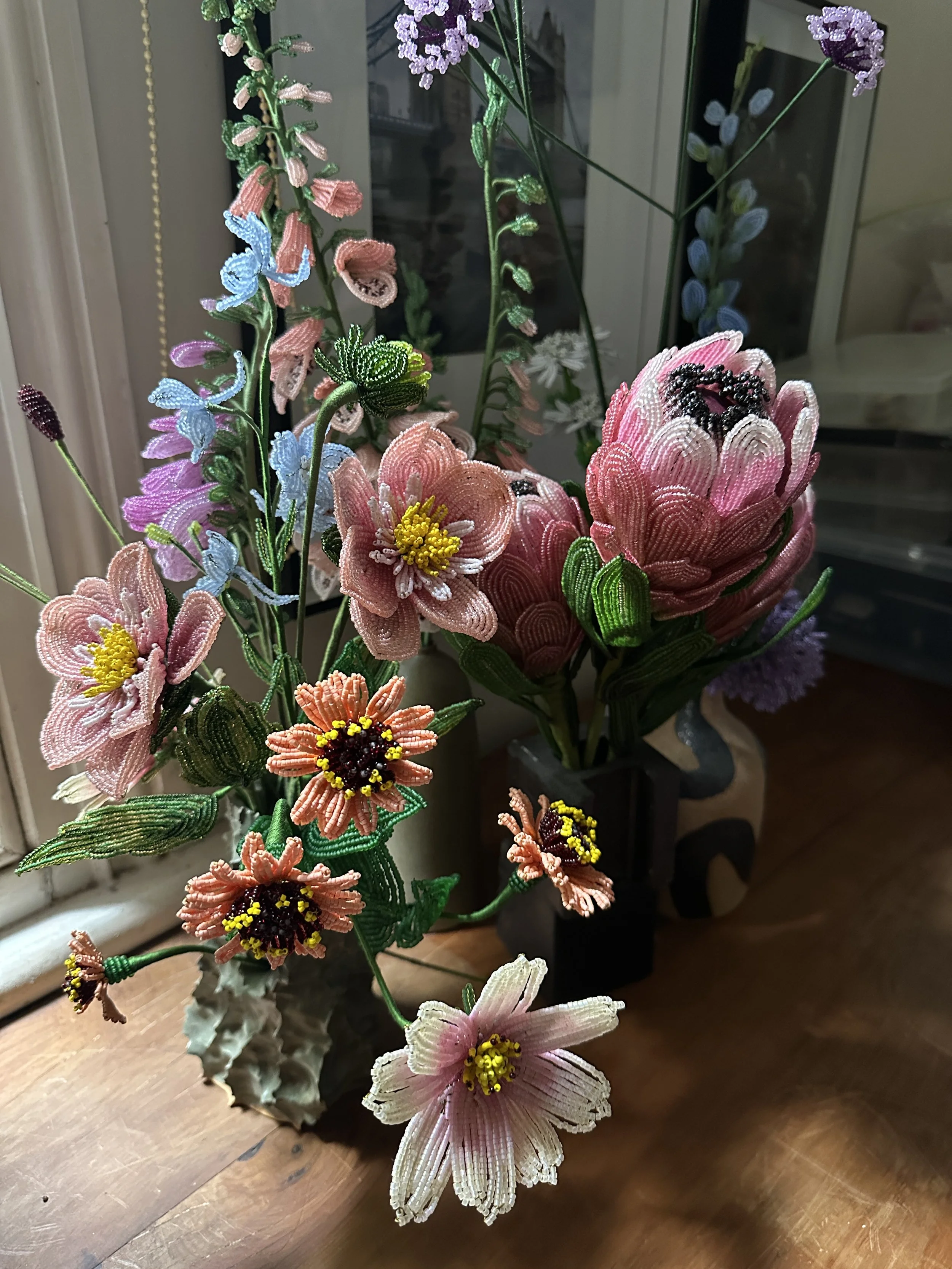 Colorful beaded floral arrangement with pink, yellow, purple, and white flowers in vases placed on a wooden table near a window.