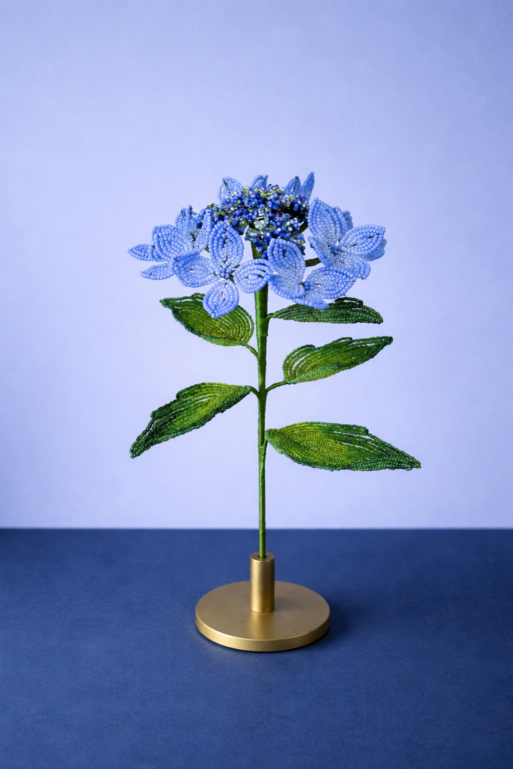 Beaded blue and green floral hydrangea sculpture on a gold base, set against a light background and dark surface by Hark Handmade