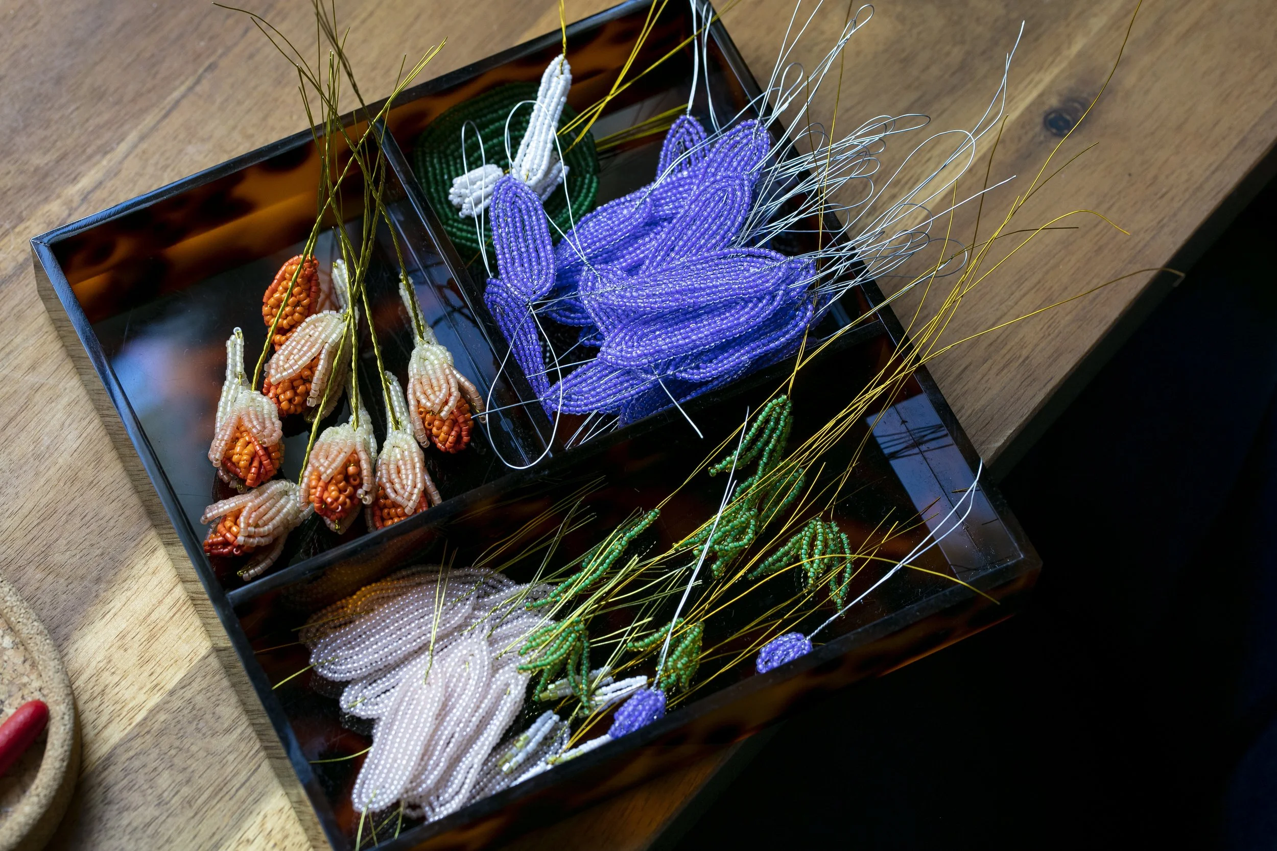 Various intricately beaded flower and leaf designs in small containers, with purple, white, orange, and green beads, displayed on a wooden surface in the Hark handmade Studio.