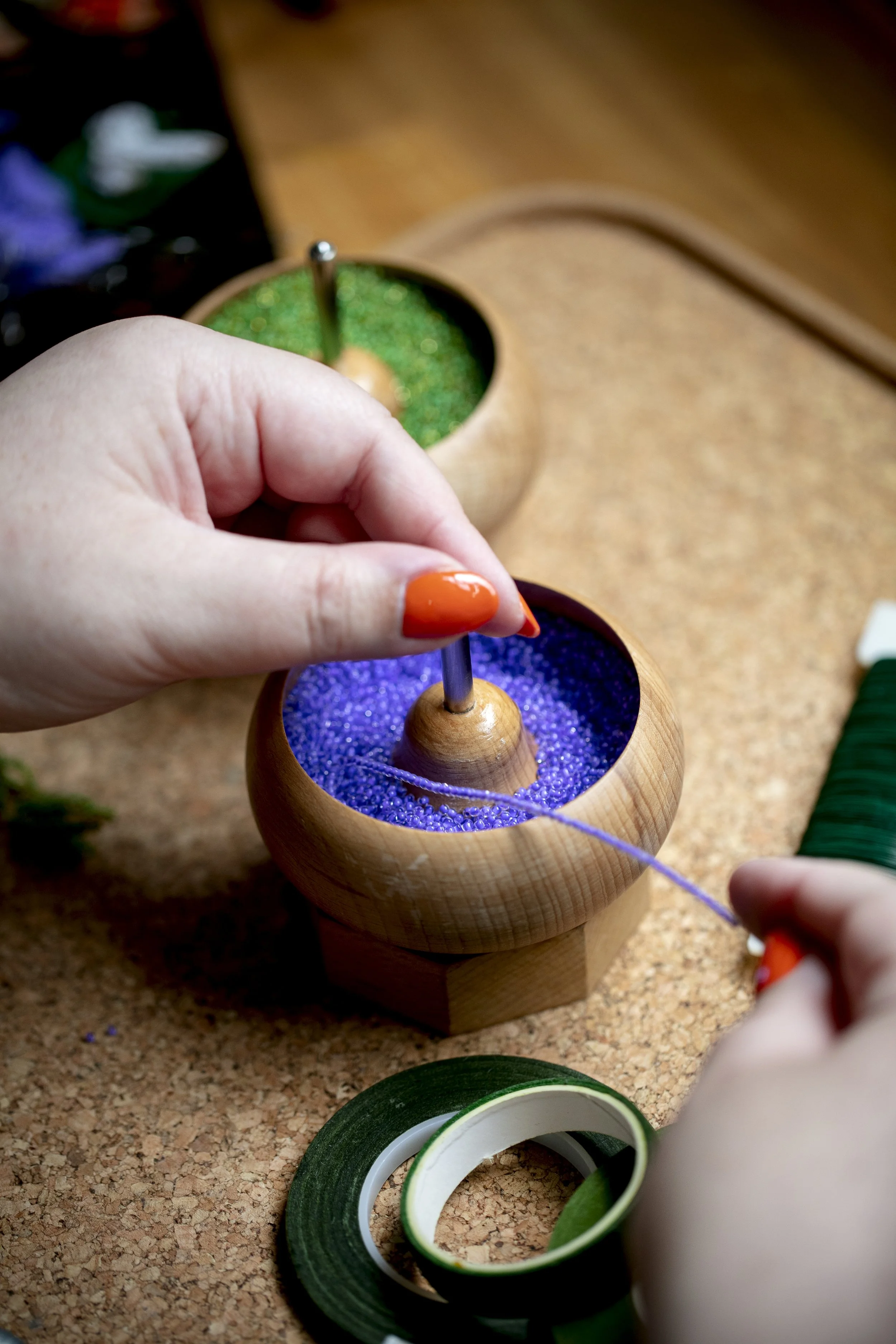 Hollie from Hark Handmade threading purple beads from a bobbin onto a string at a beading workstation with various bead containers and green tape nearby.