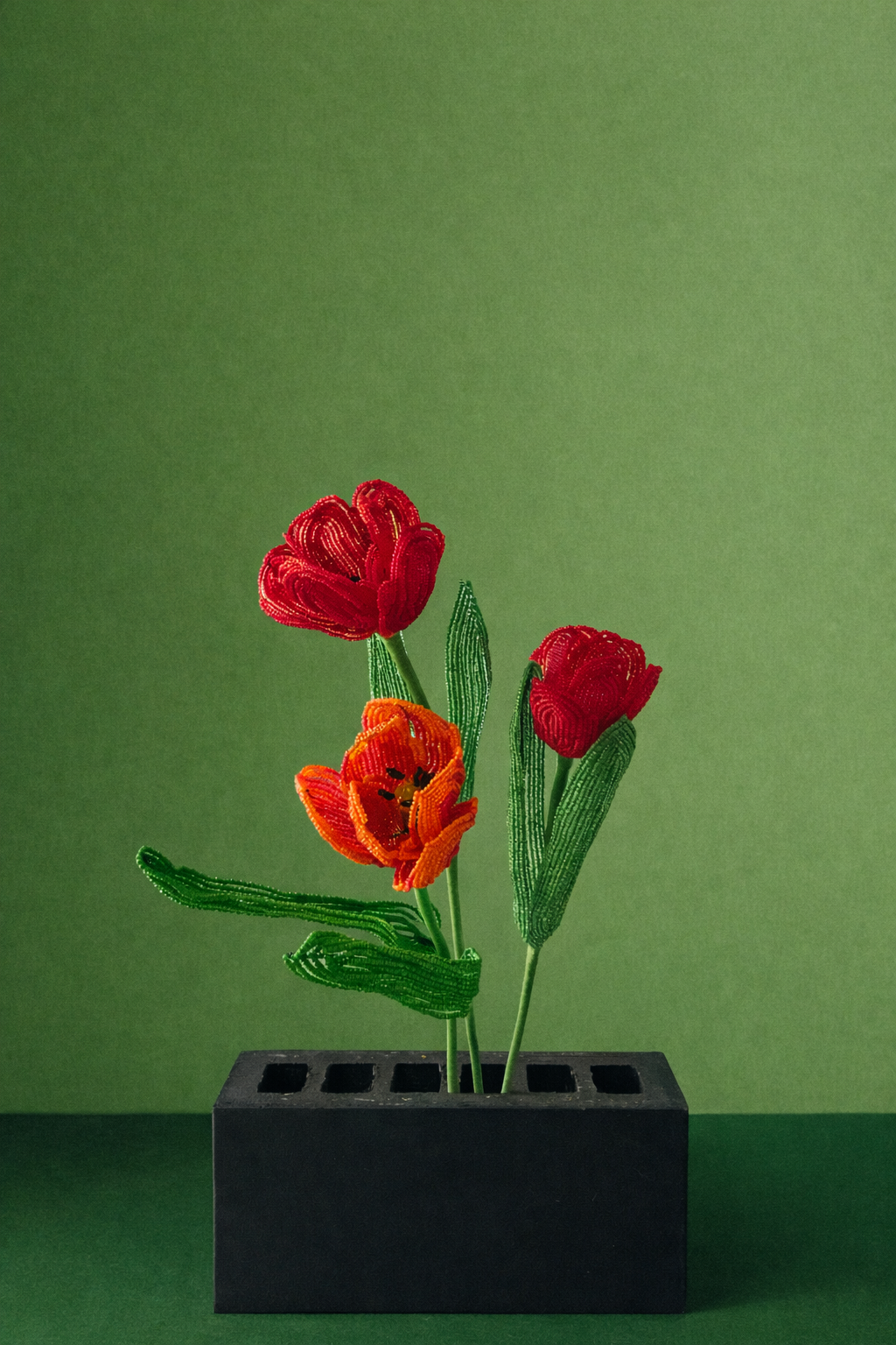 Beaded flower arrangement with red, orange, and red flowers in a black rectangular container, against a green background.