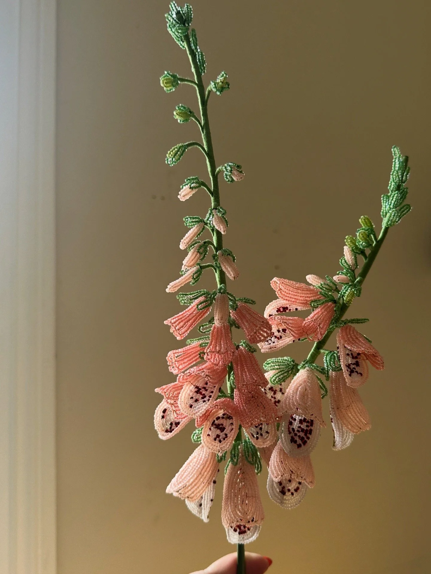 A beaded flower crafted with pink and green beads, resembling a bellflower with multiple blooms hanging from a central green stem, against a beige background.