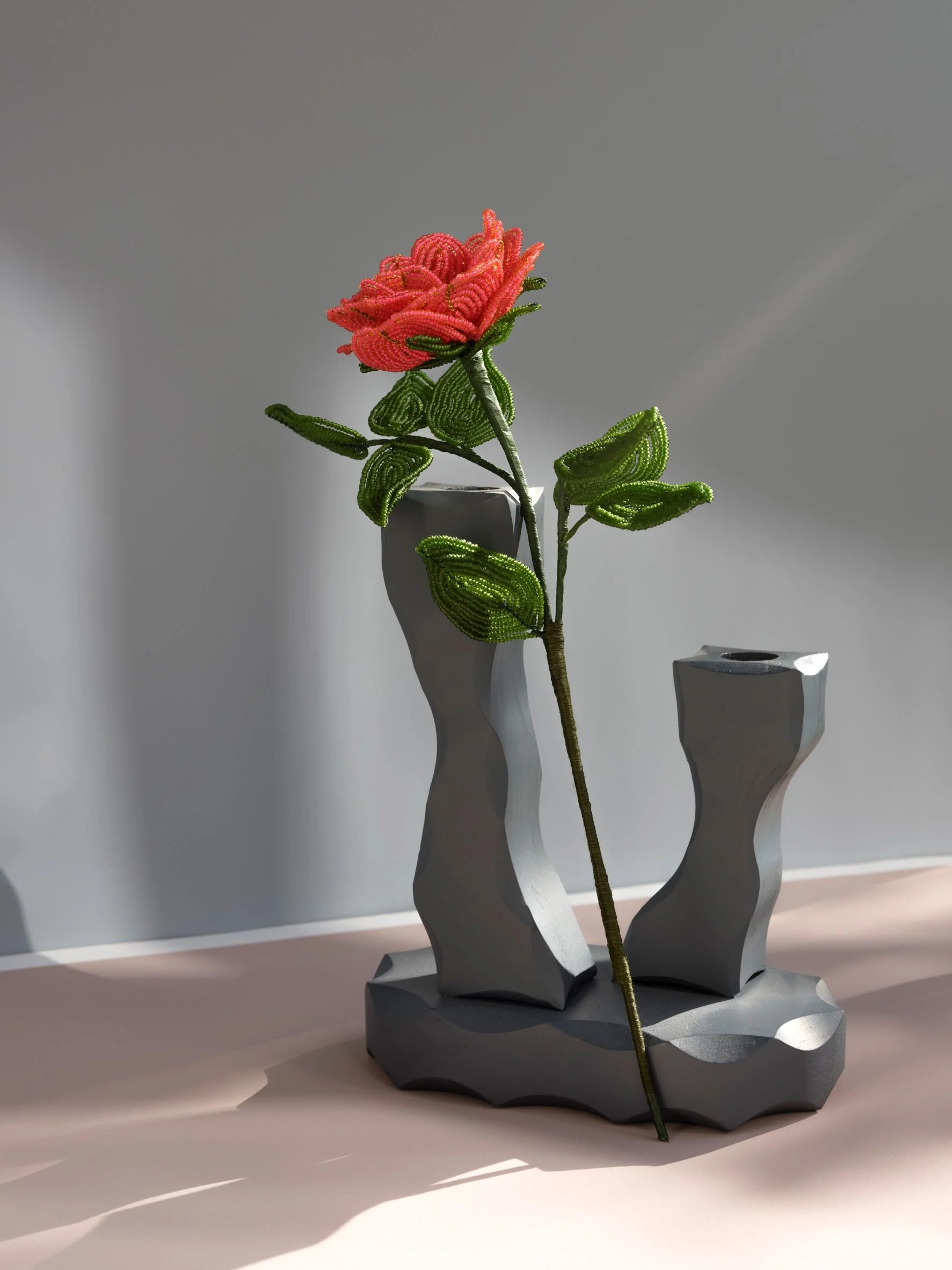 Beaded pink and green flower in a modern gray vase by Hark Handmade Beaded Botanical Sculptures, with abstract, textured shapes on a pink surface with a light gray background.