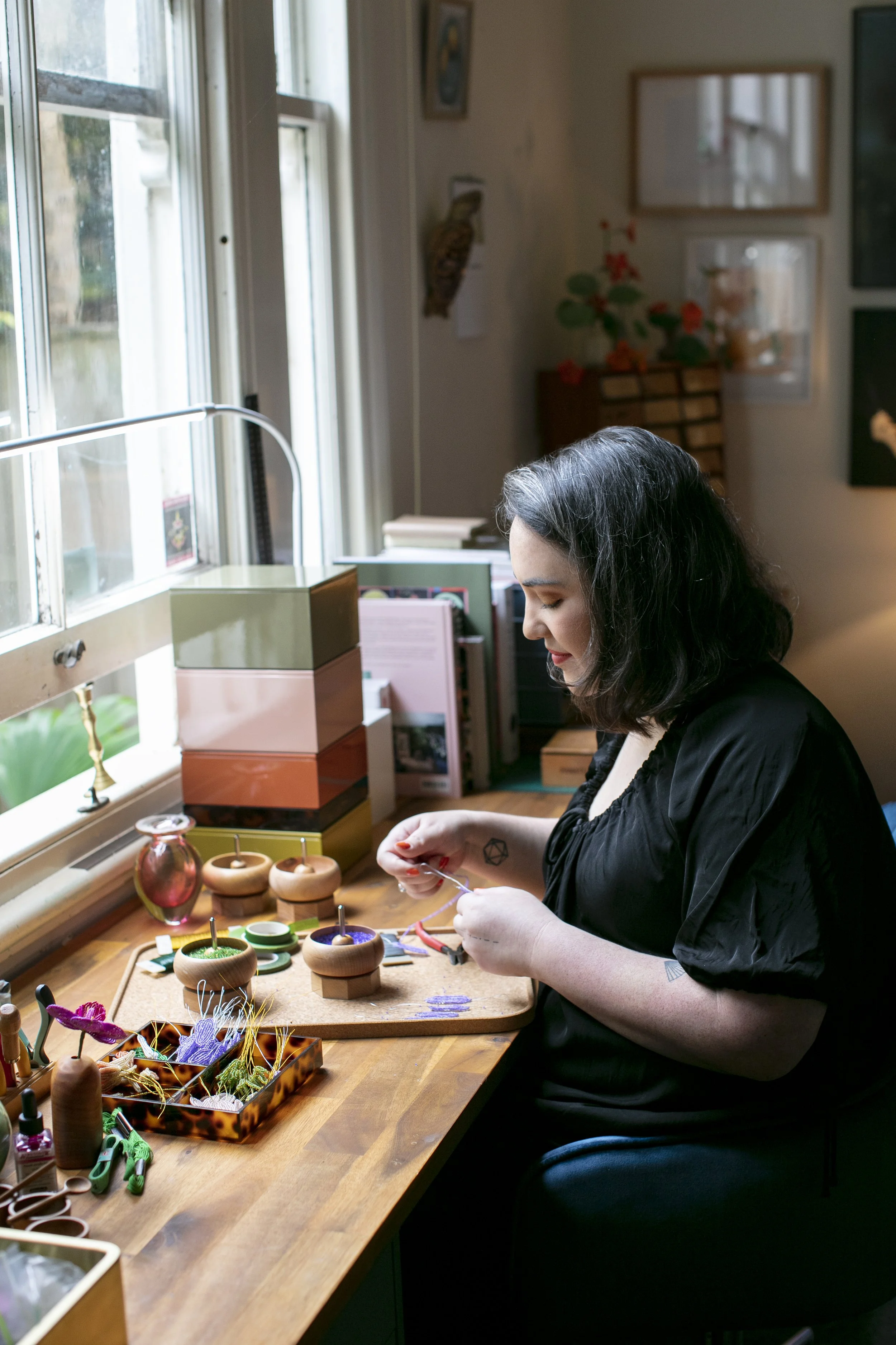 A woman (Hollie Ryan of Hark Handmade) with shoulder-length black hair sitting at a wooden desk by a window, engaged in hand beaded flower making.