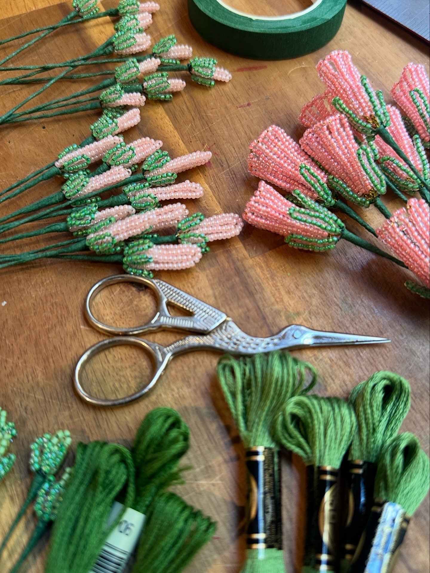 Crafting supplies including pink and green beaded flower stems, green floral tape, and green floral wire, along with a pair of small scissors on a wooden surface.