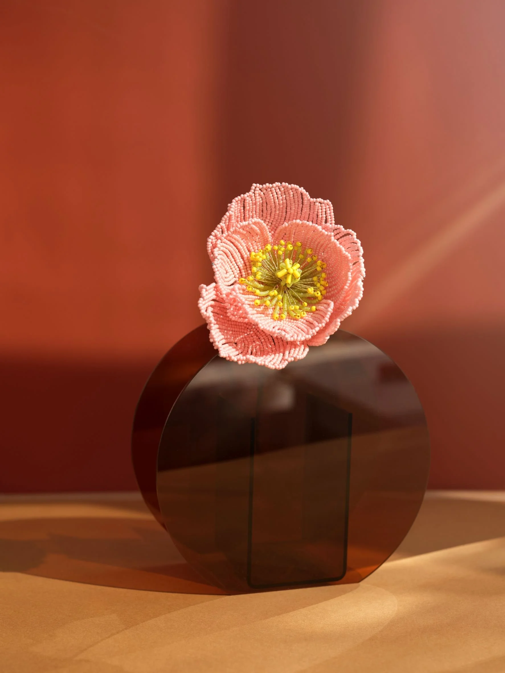 A pink beaded flower sculpture with yellow centers, mounted on a glossy brown circular stand, against a pink background. Photo taken by Lula Cucchiara for Homestyle Magazine.
