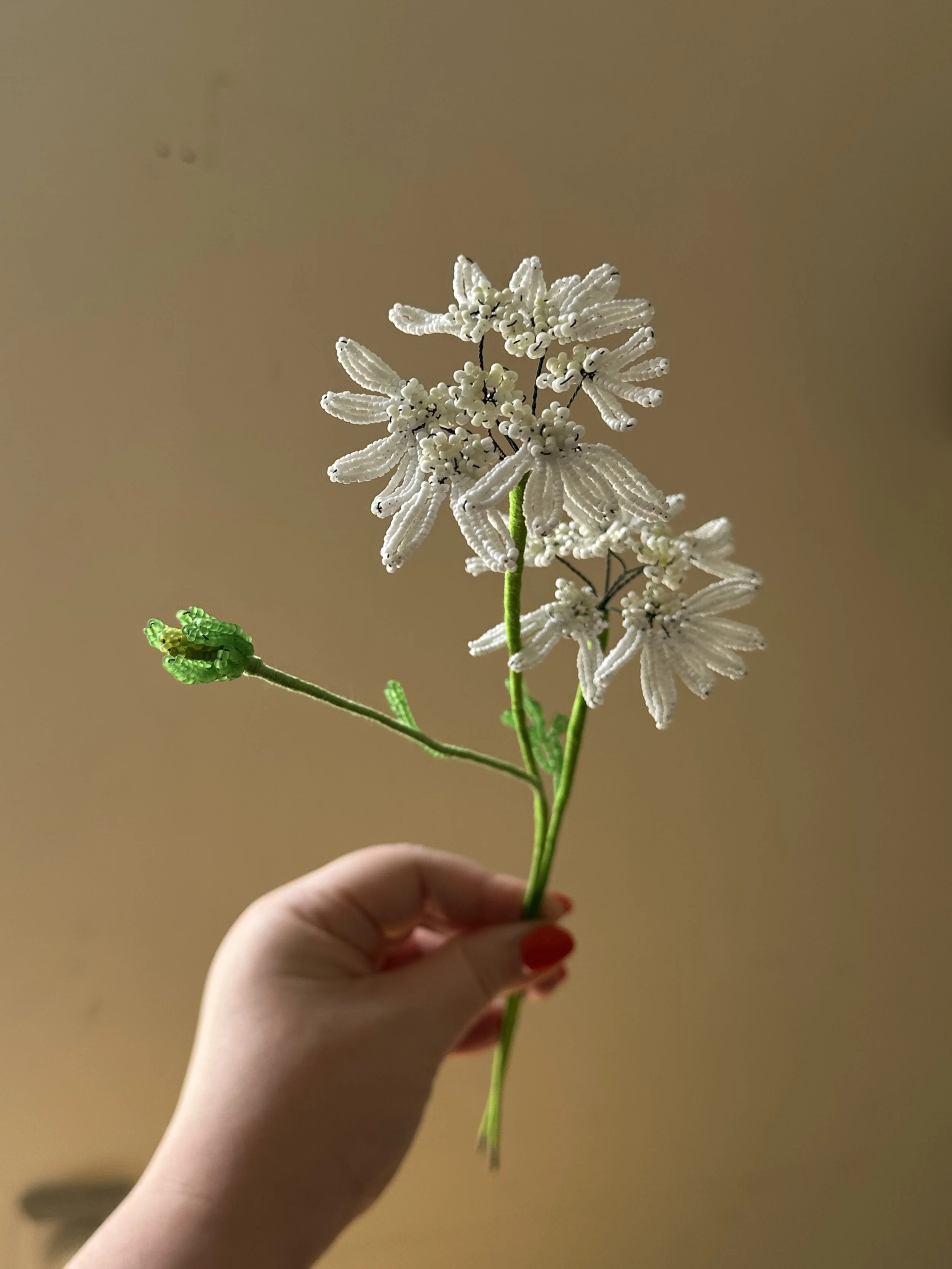 Hand holding a beaded floral craft resembling white flowers with green leaves on a neutral background.