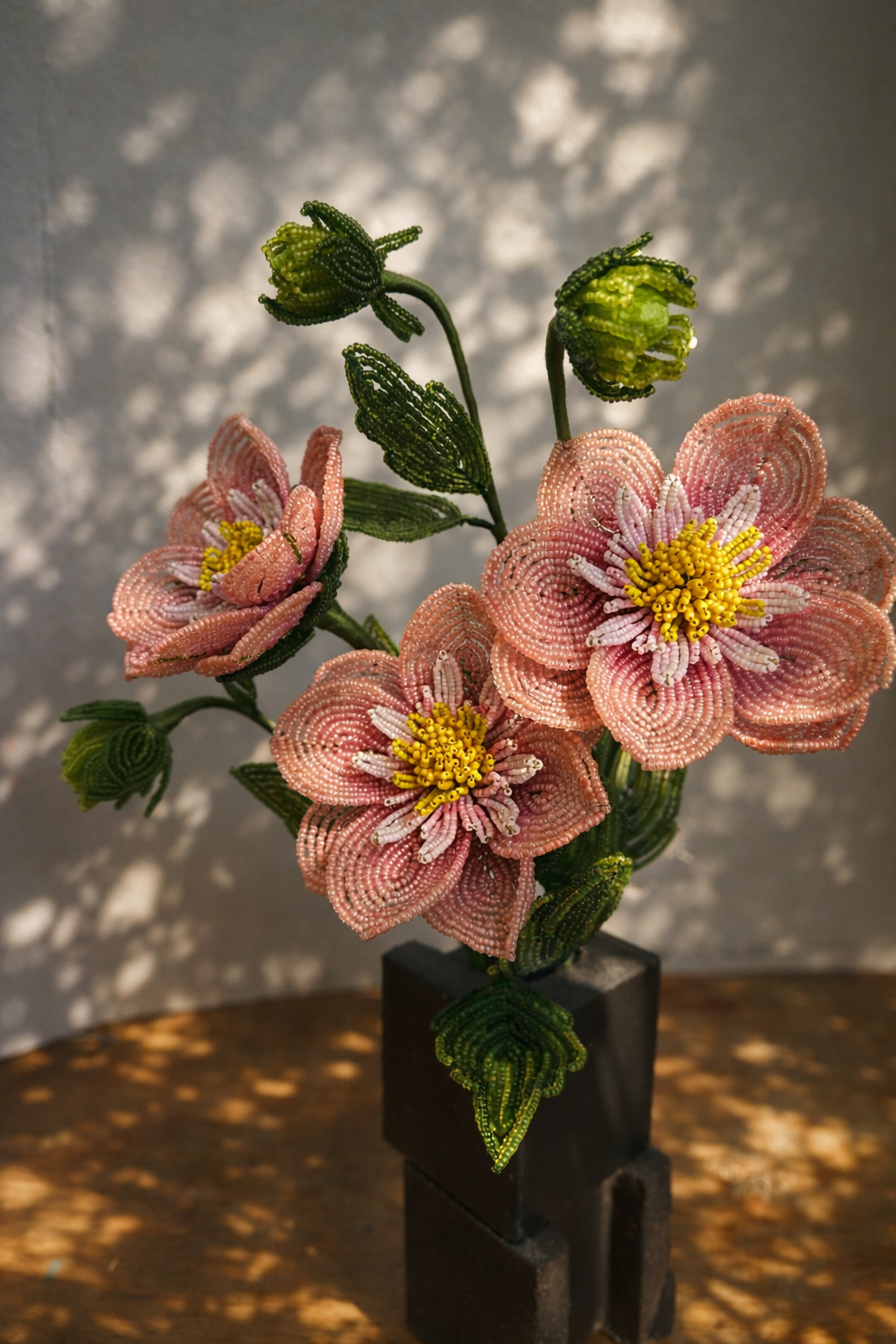 Beaded flower sculpture of pink flowers with yellow centers and green leaves, in a black square vase, on a wooden surface with patterned shadows by Hark Handmade