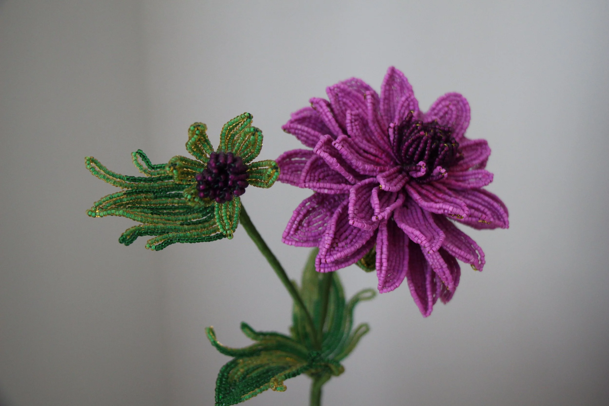 Beaded craft flower with purple petals and green leaves, crafted from small beads.
