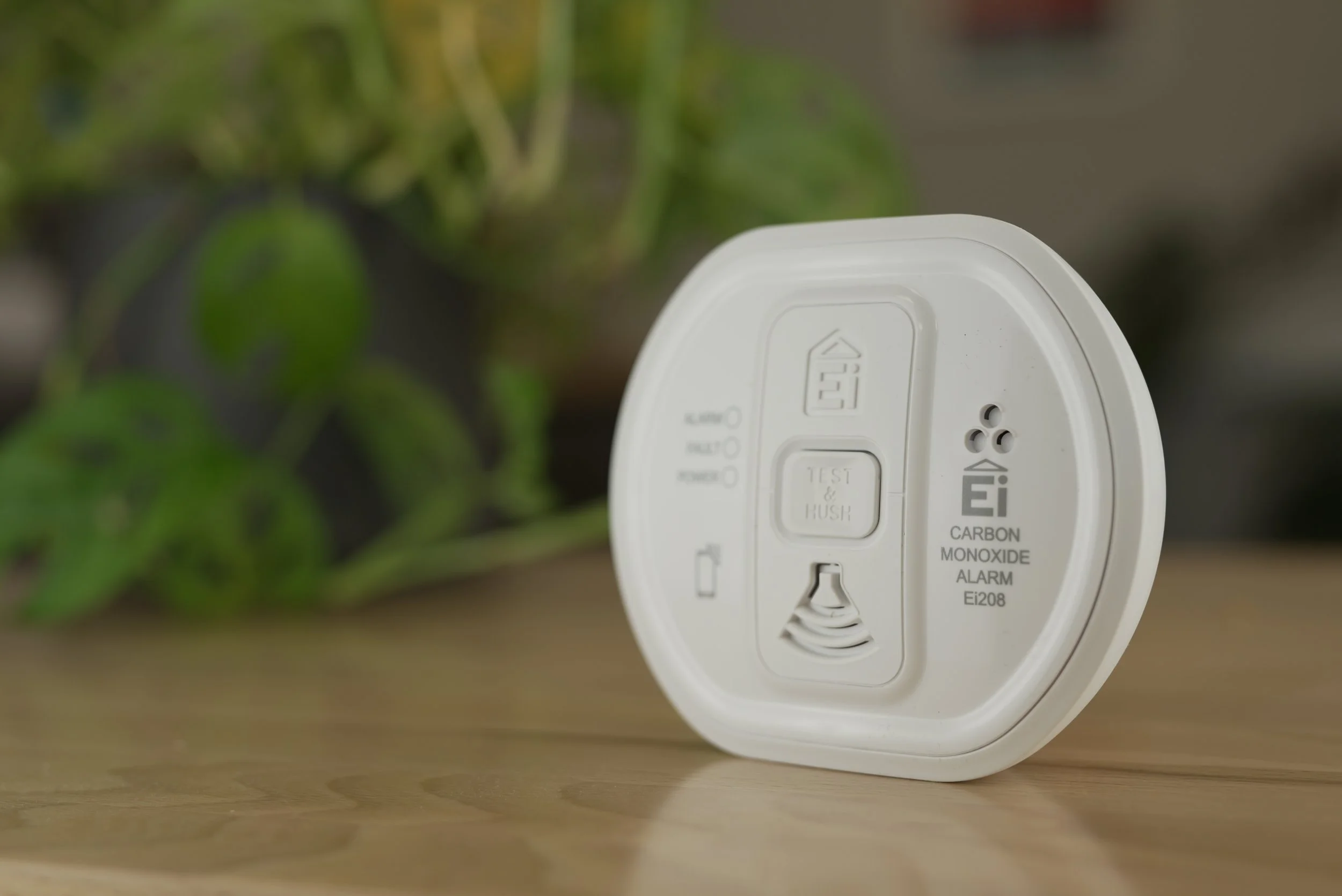 A white carbon monoxide alarm on a wooden surface with green plants in the blurred background.