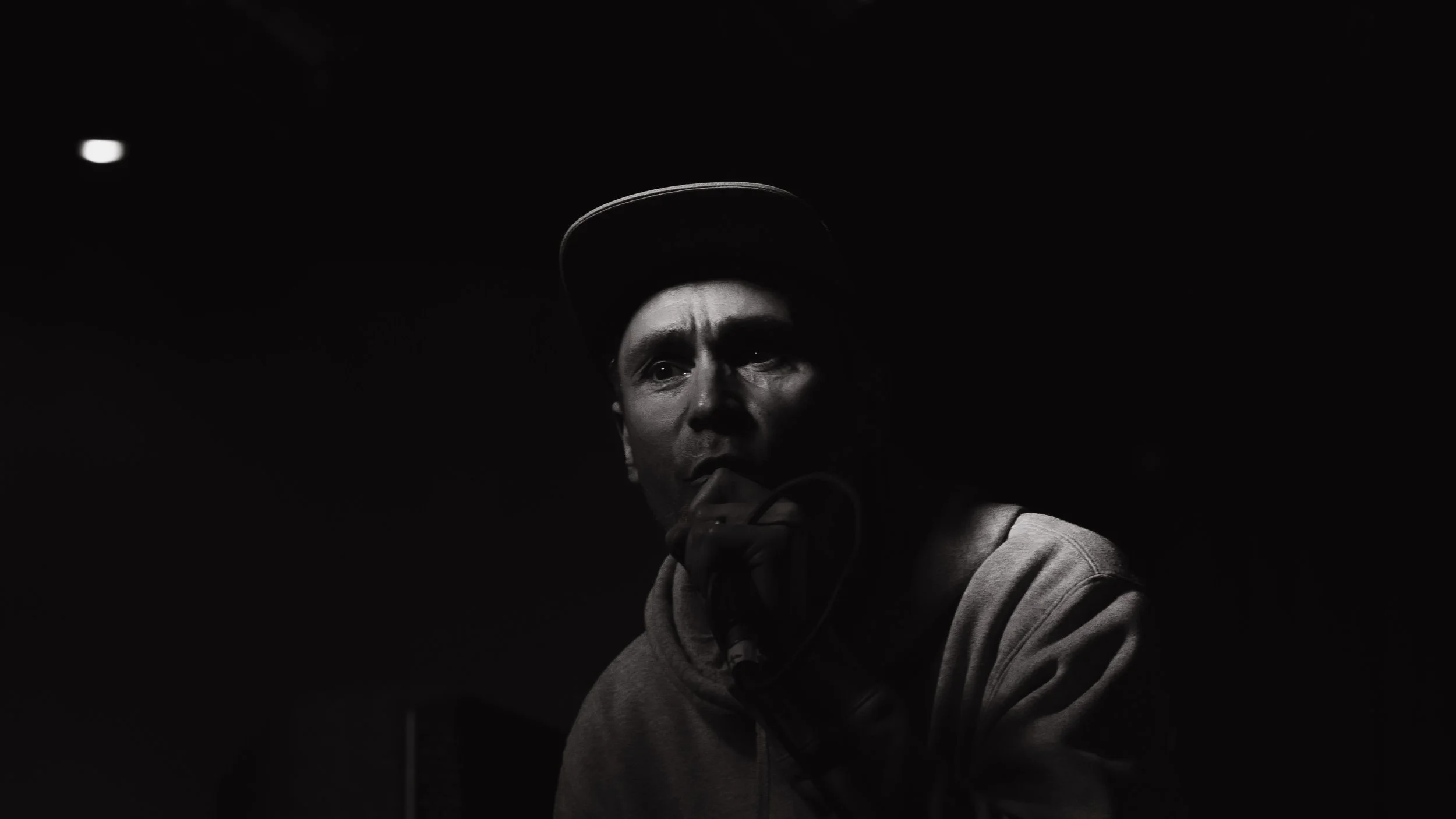 A black and white photo of a man wearing a hoodie and a cap, holding a microphone close to his mouth, with a serious expression and shadows on his face.