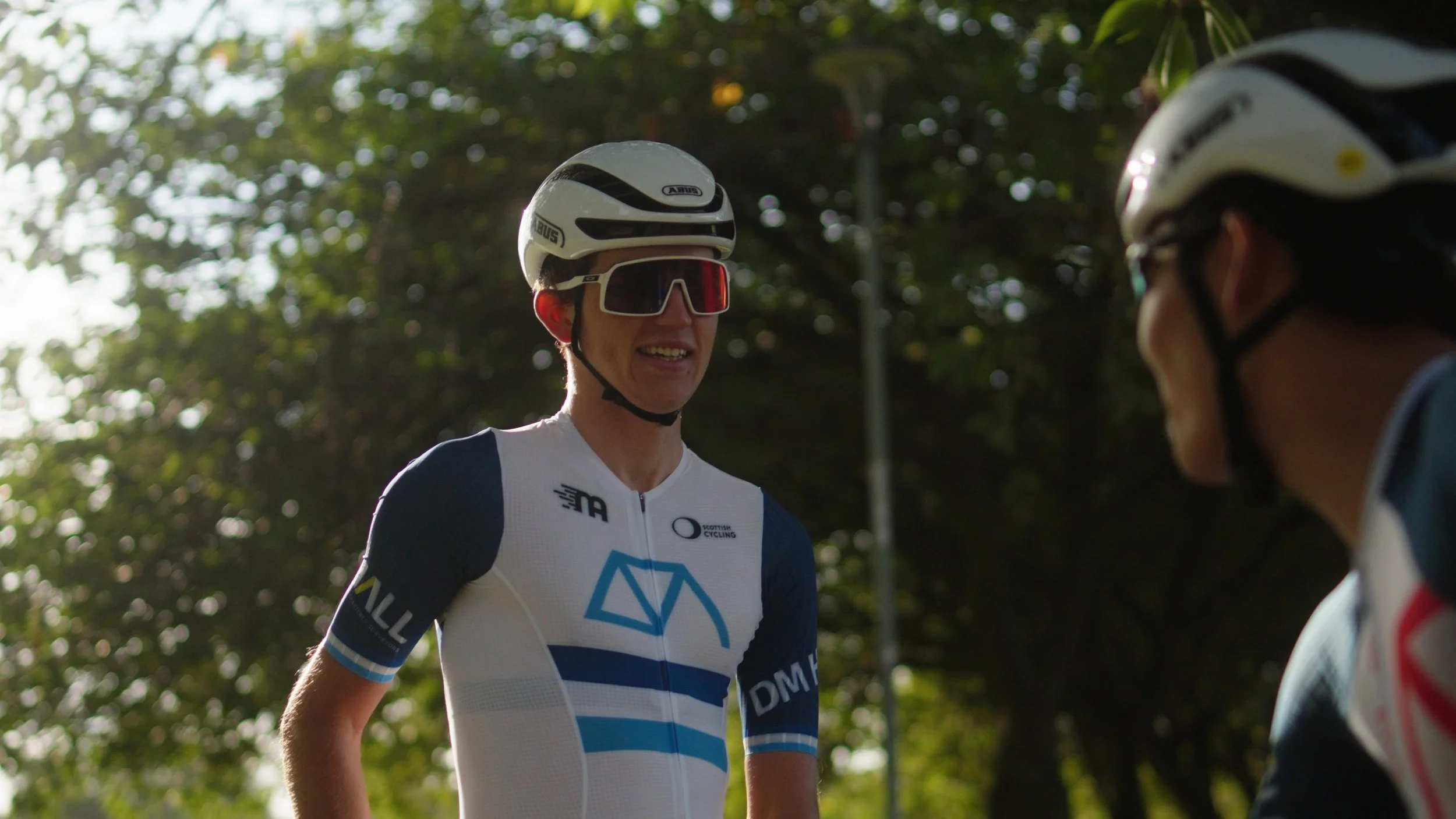 Two cyclists wearing helmets and sports jerseys are having a conversation outdoors with trees in the background. One cyclist is smiling.