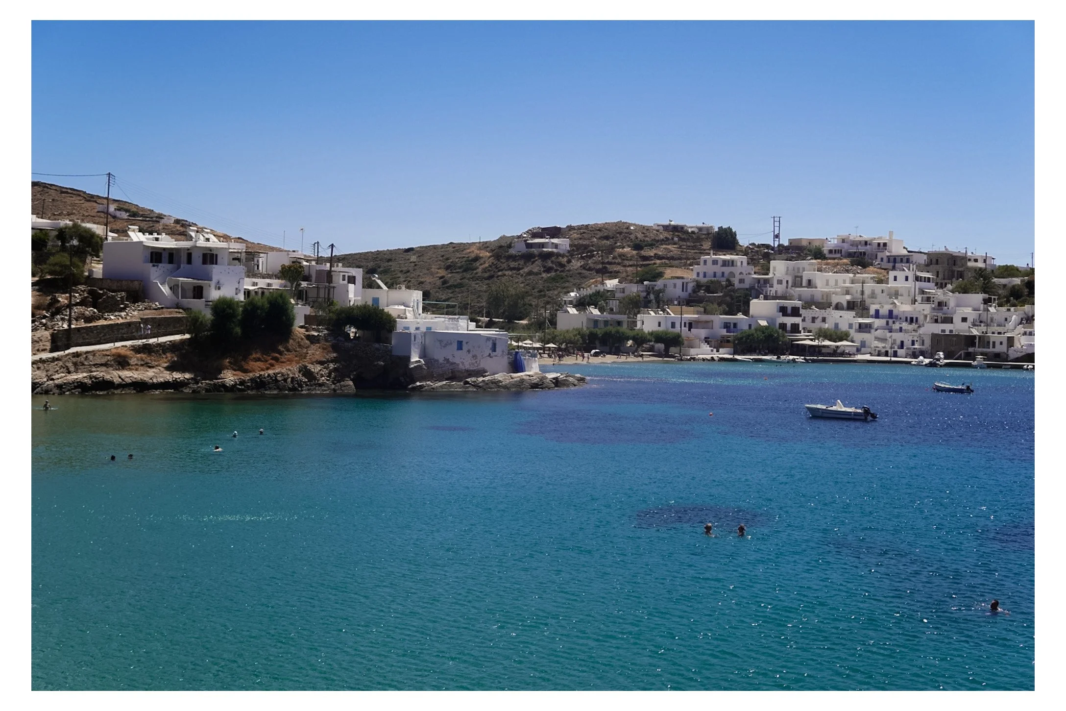 A coastal scene with turquoise water, white buildings on hills, and a clear blue sky, with some people swimming and boats floating in the water.