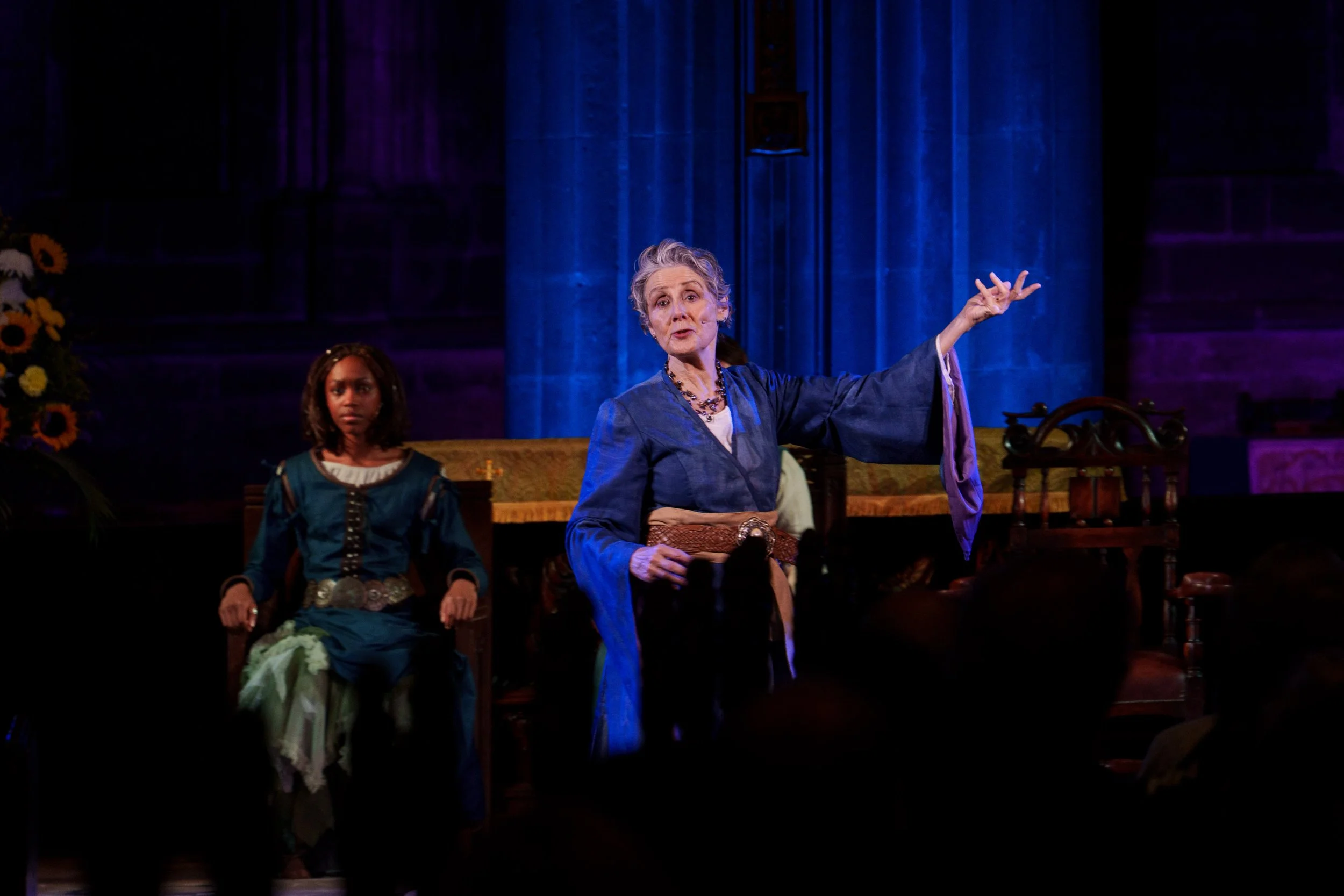 An elderly woman dressed in a blue costume performs on stage with her hand gesturing outward, while a young girl watching stands behind her. The scene is set in a dimly lit theater with a dark background.