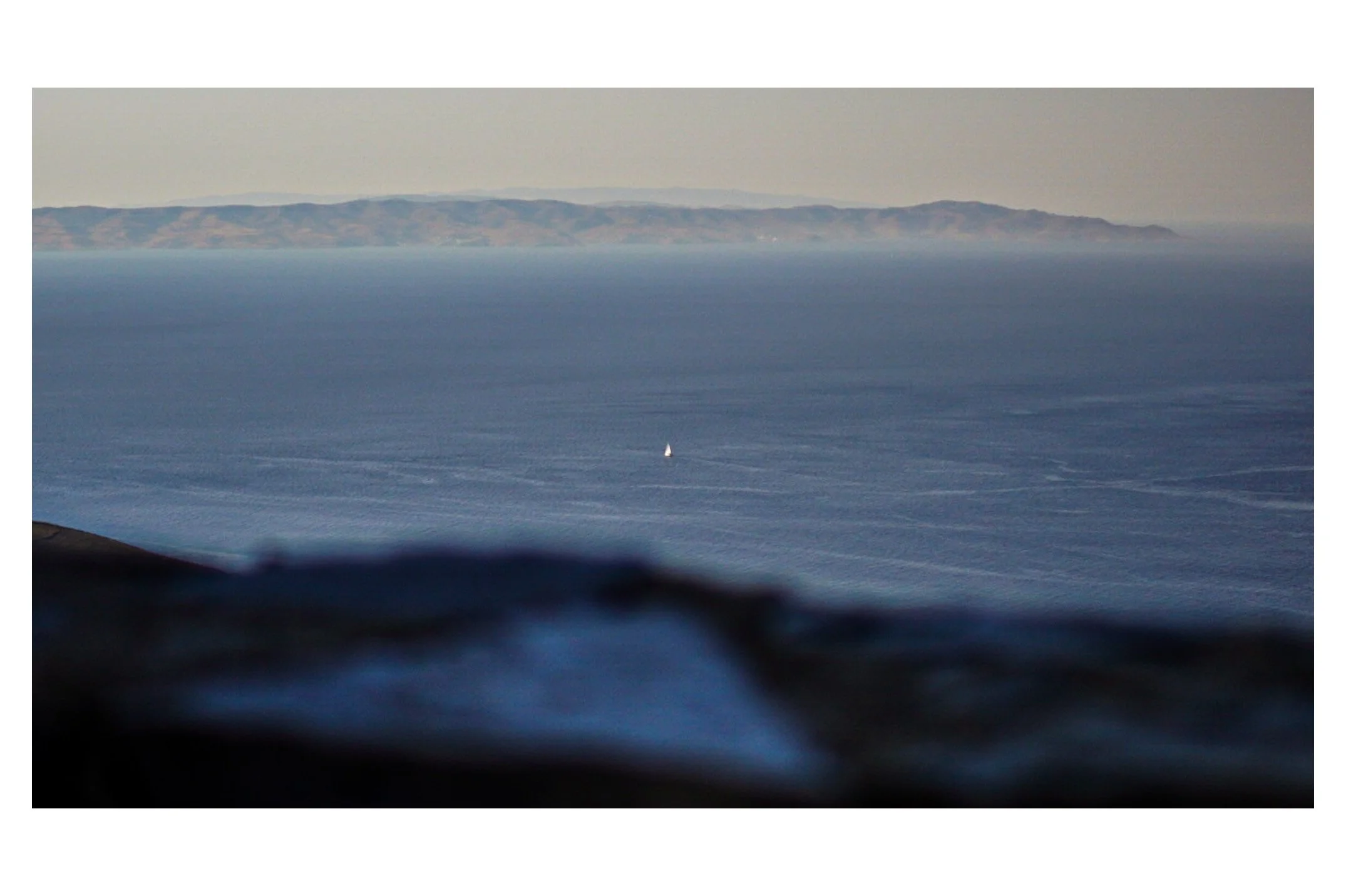 Open ocean with a small sailboat, distant landforms in the background, and a blurry foreground.