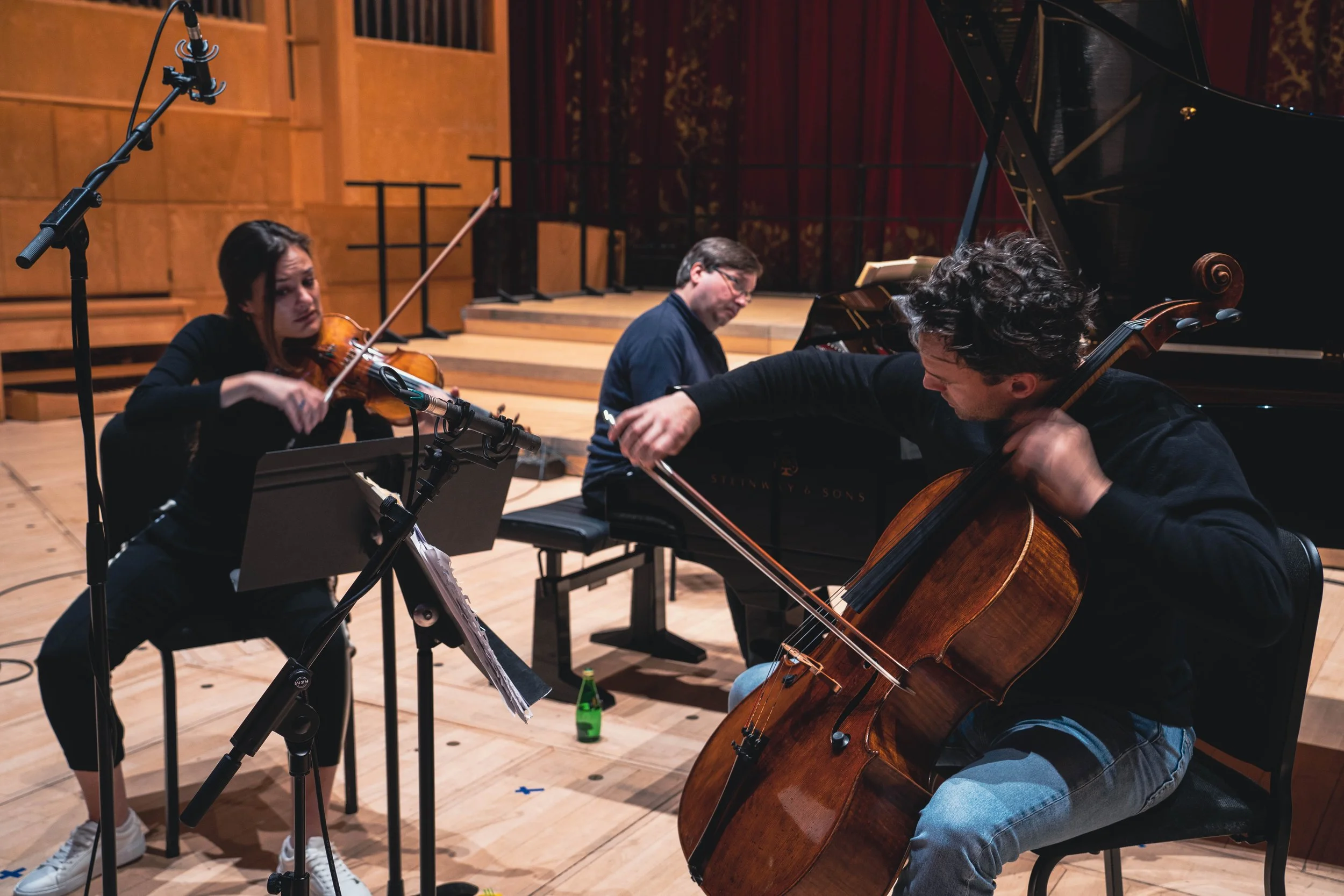 Three musicians rehearsing in a concert hall: a violinist on the left, a pianist in the center, and a cellist on the right.