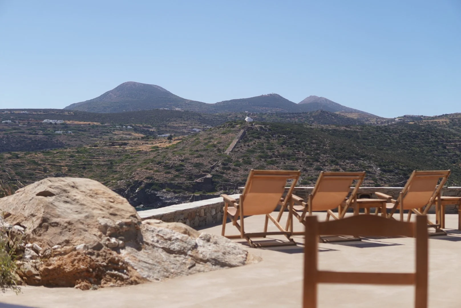 Three wooden deck chairs on a patio overlooking a hilly landscape with mountains in the background on a clear sunny day.