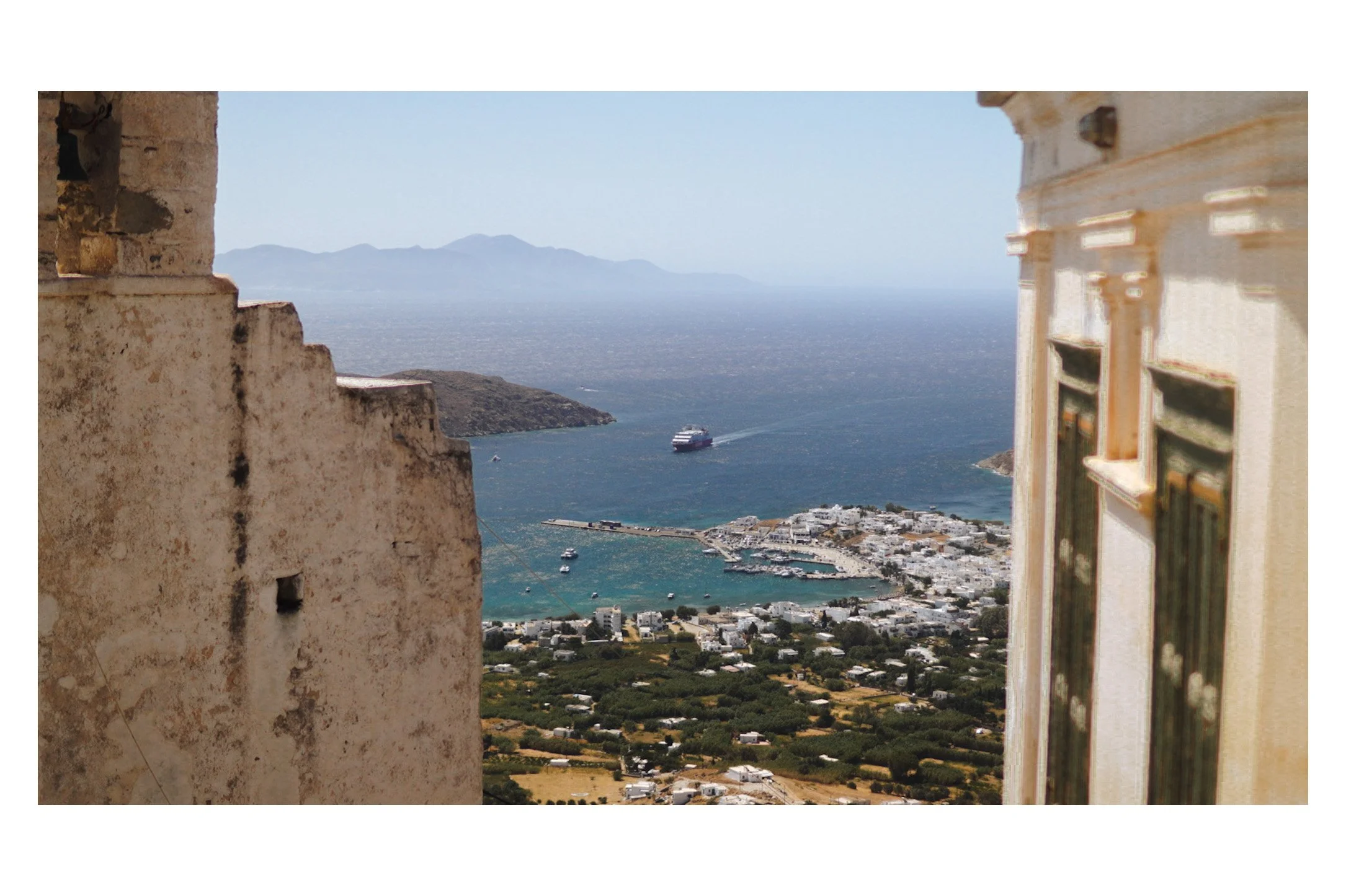 View of a coastal town with boats, a cruise ship in the water, and mountains in the distance, framed by two old buildings.
