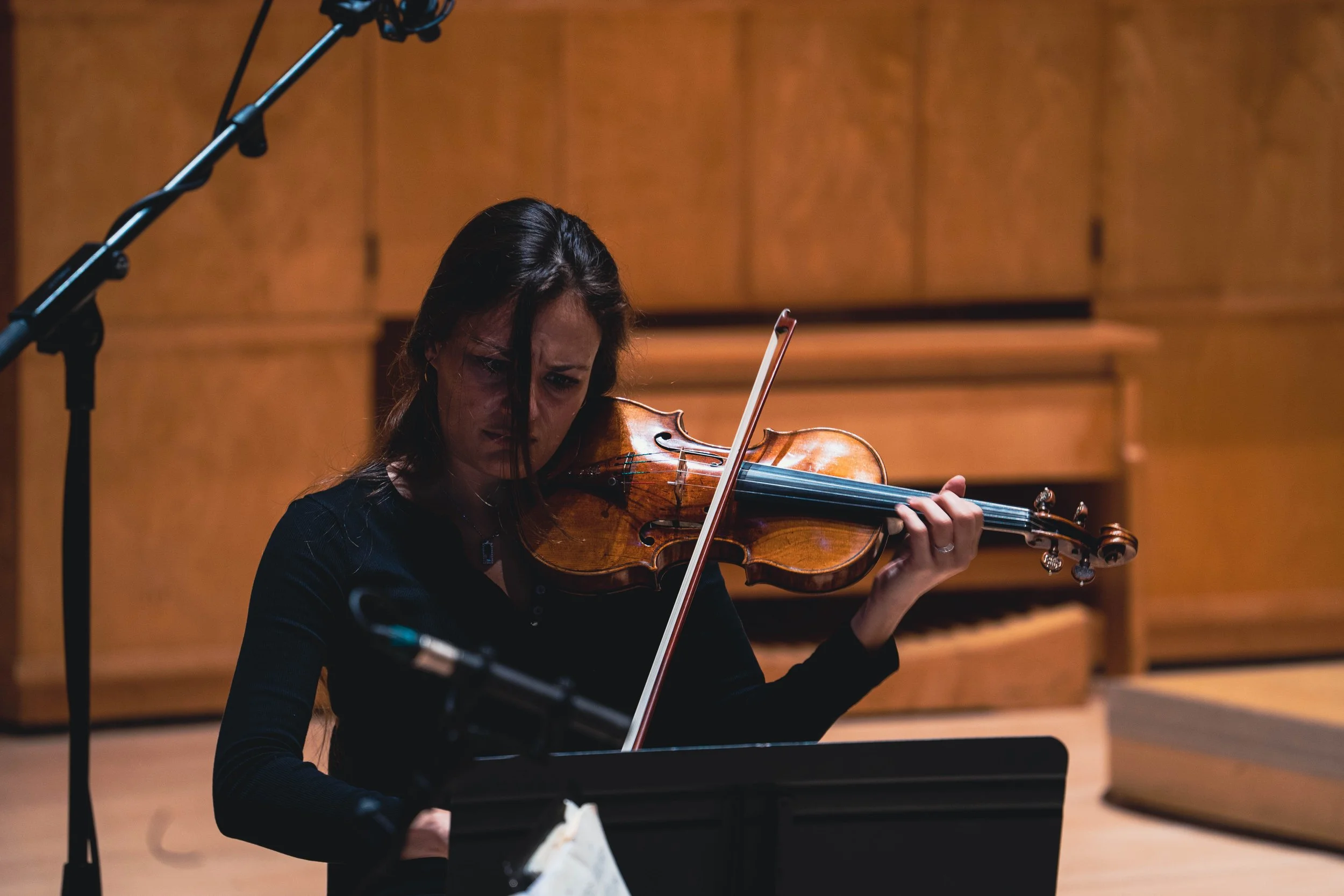 A woman playing the violin in a concert hall with a wooden background, with microphones nearby.