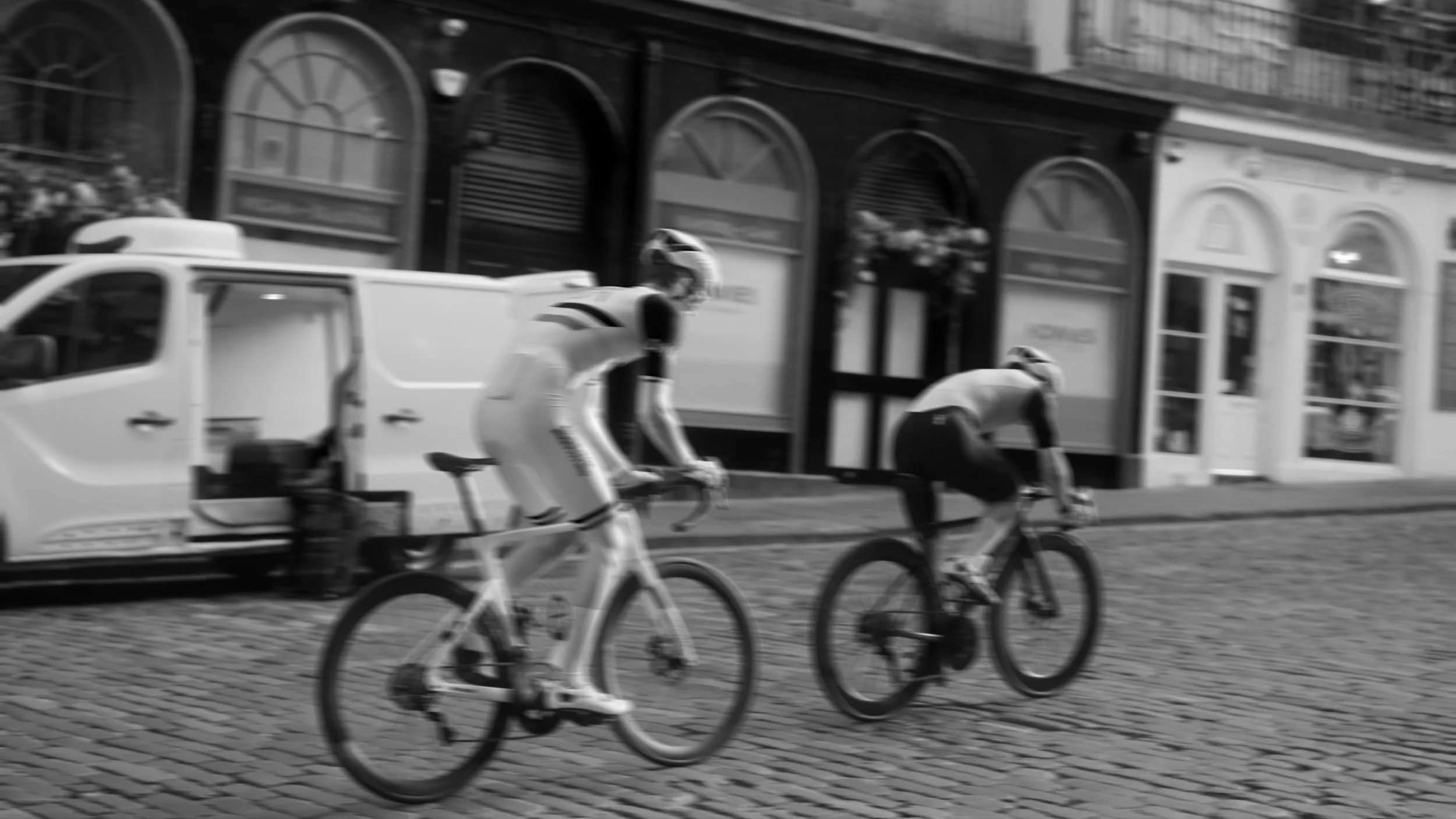 Two cyclists riding on a cobblestone street in front of a row of shops and parked vehicles in an urban setting.