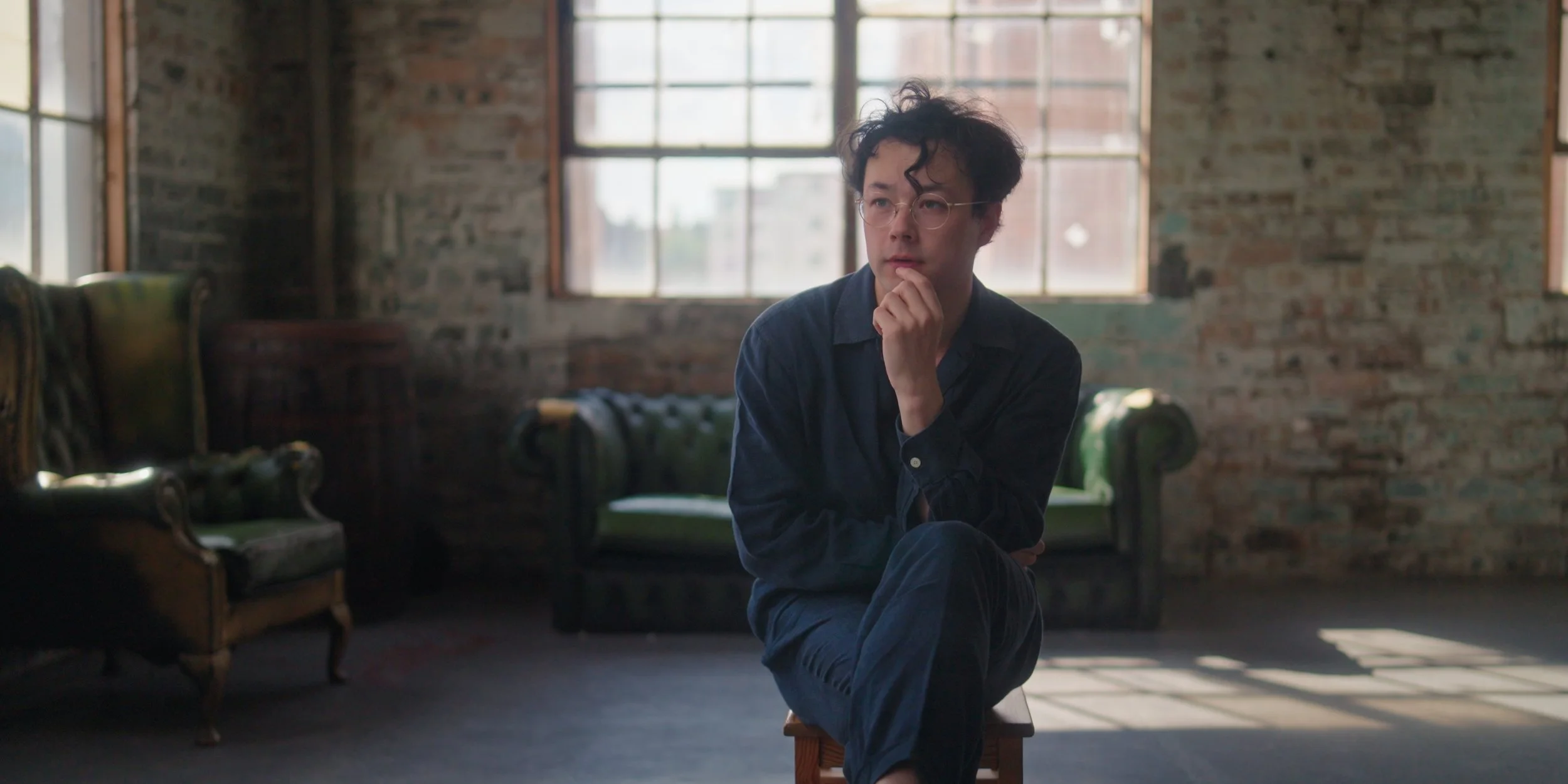 A man sitting on a small stool with his chin resting on his hand, inside a room with exposed brick walls and large windows.