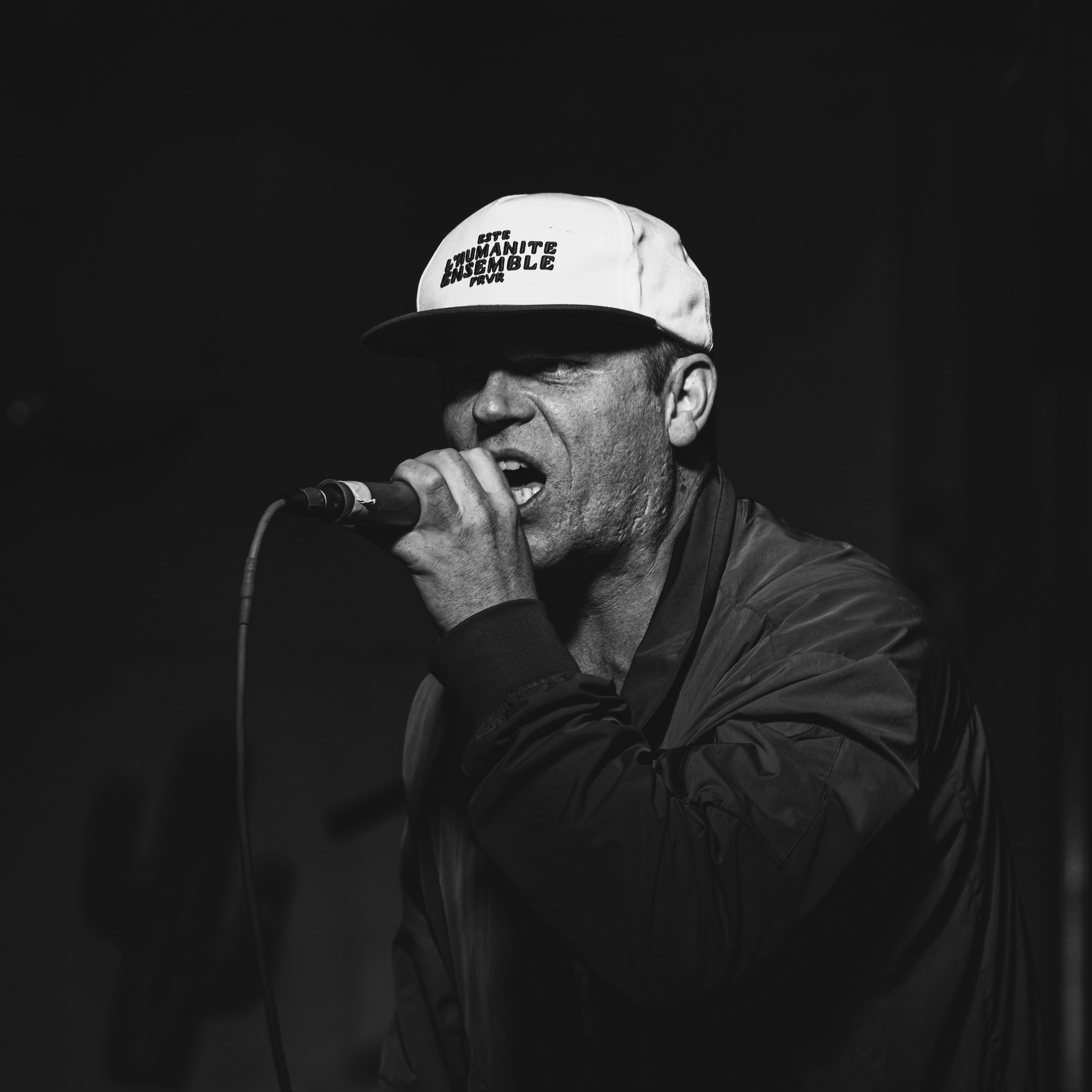 A man singing into a microphone, wearing a white cap with text and a dark jacket, in a black and white photo.
