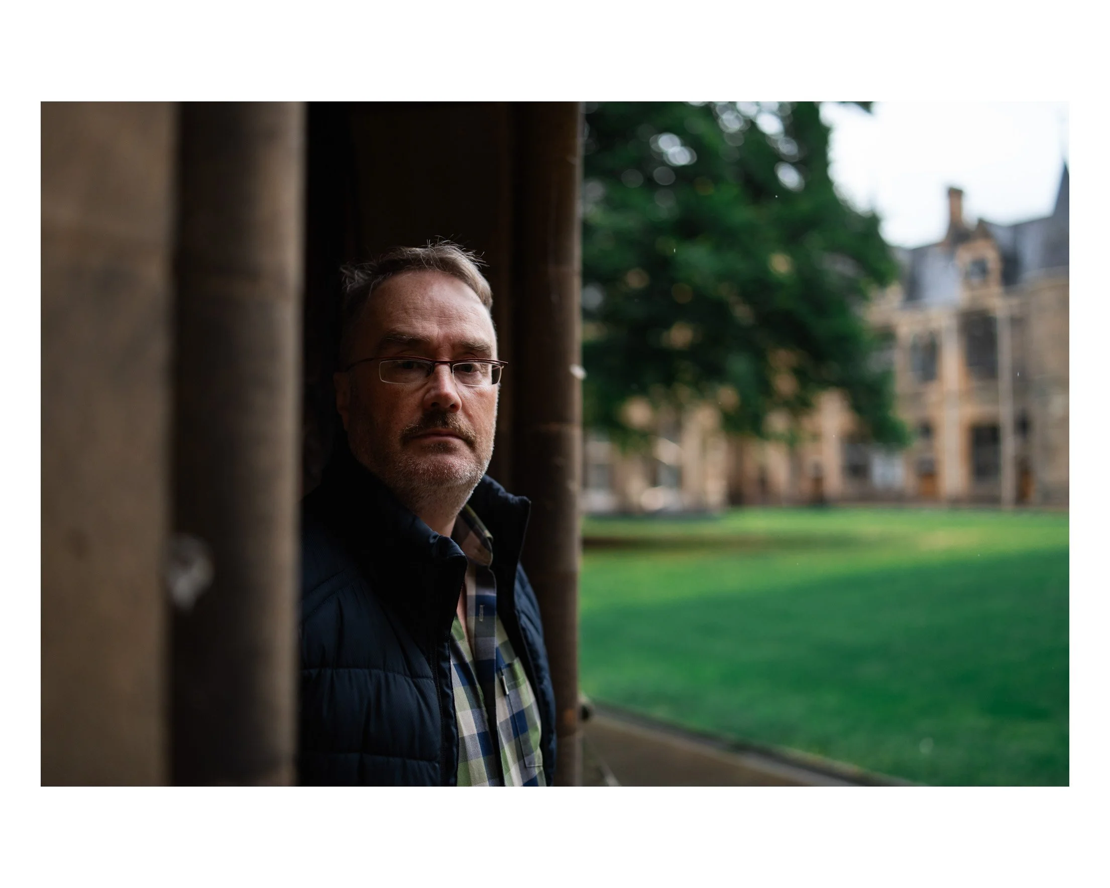 A man with glasses stands outdoors, partly hidden behind a wooden column, with a historic building and green lawn in the background.