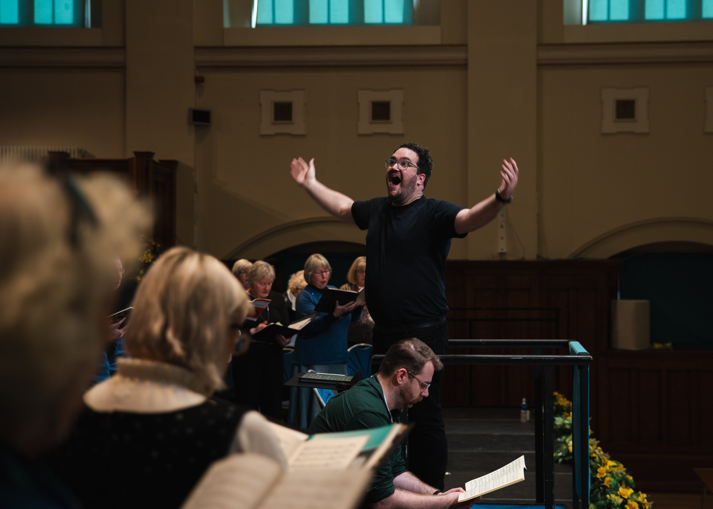 A man with glasses and a black shirt conducting a choir during a performance, with choir members singing behind him and holding sheet music.