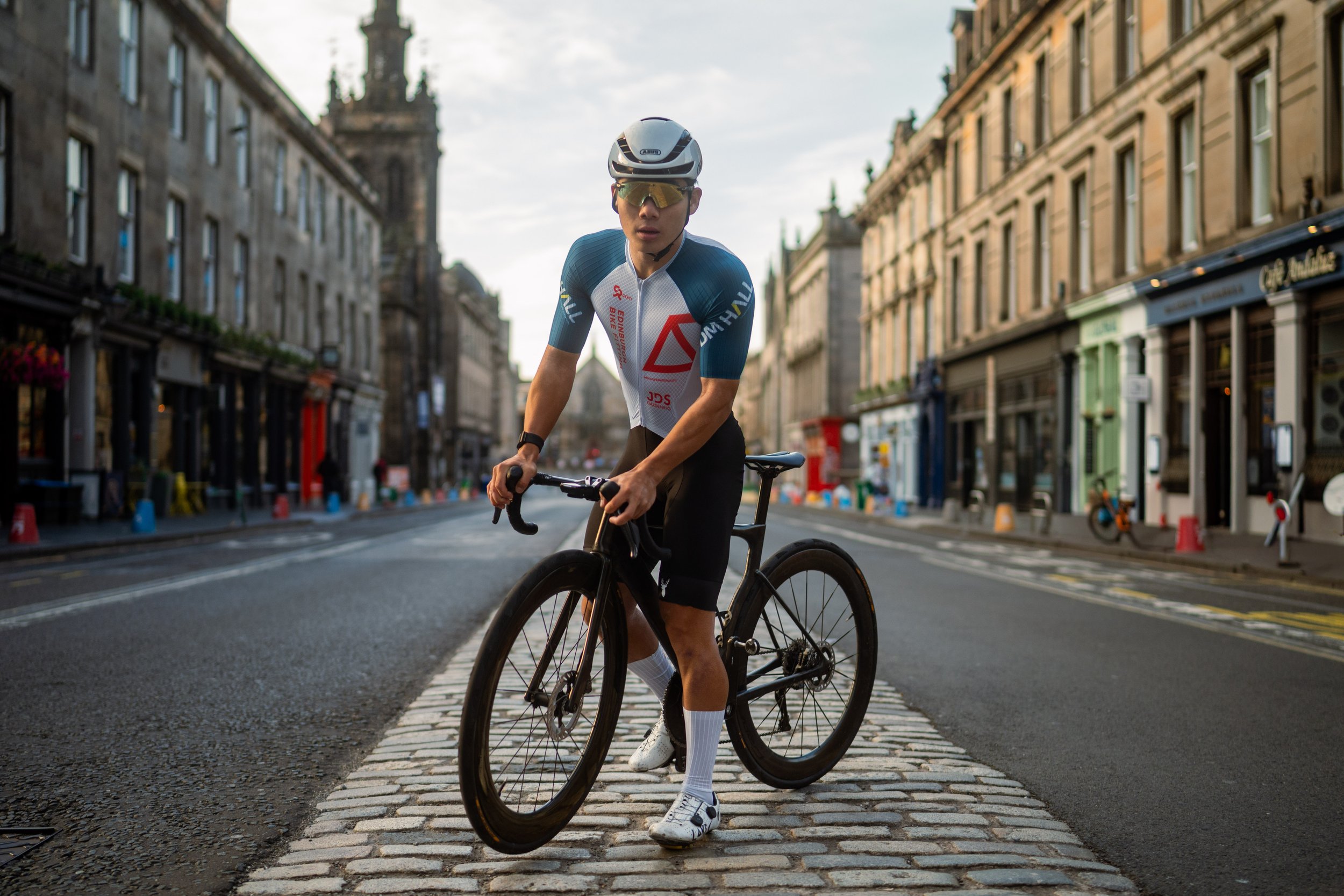 A person wearing a white helmet, sunglasses, a cycling jersey, and shorts riding a black road bike on a city street during daytime. The street is lined with historic buildings, some shops, and a church spire visible in the background.