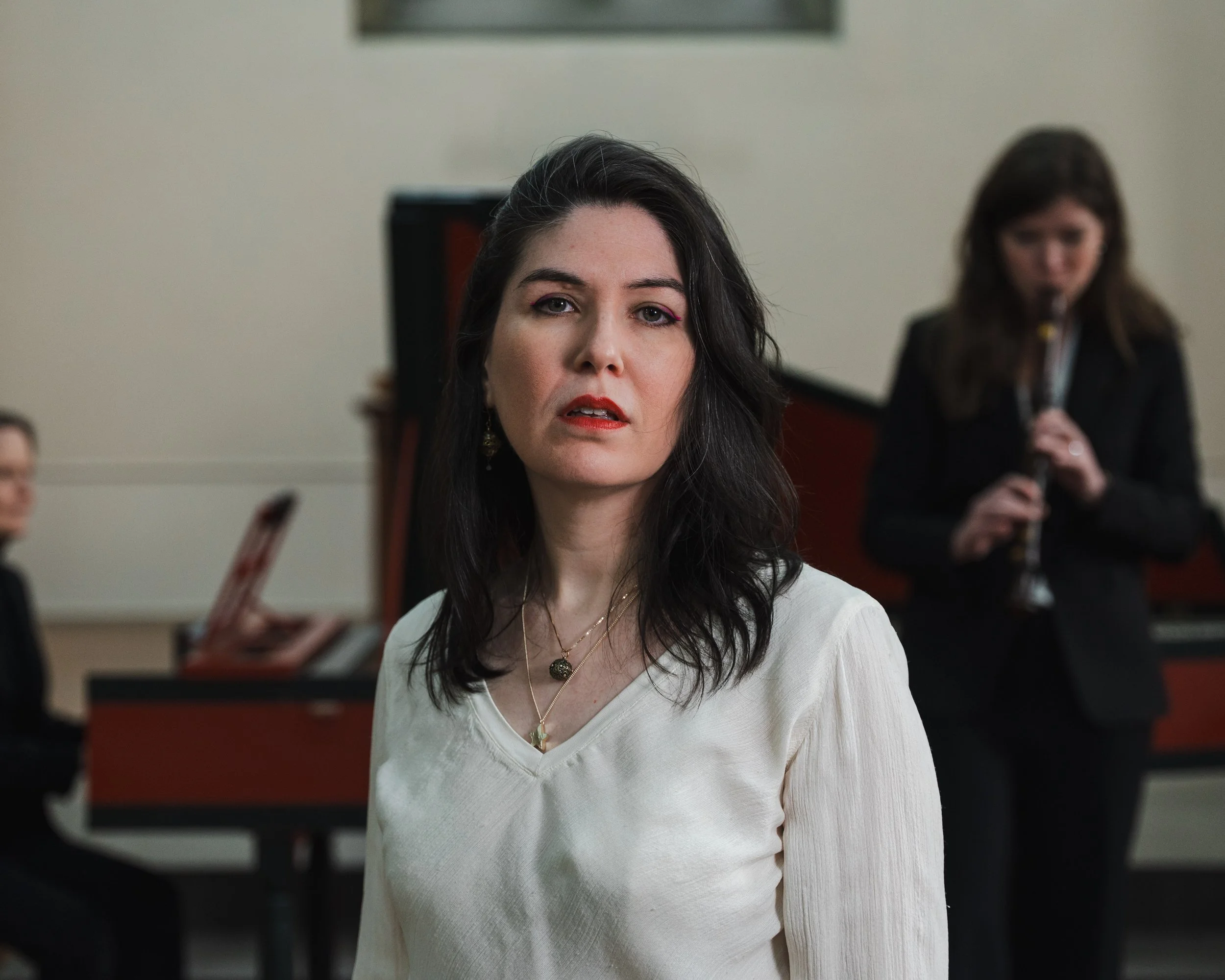 A woman with black hair in a white top looking at the camera, with a pianist and a woman playing the flute in the background