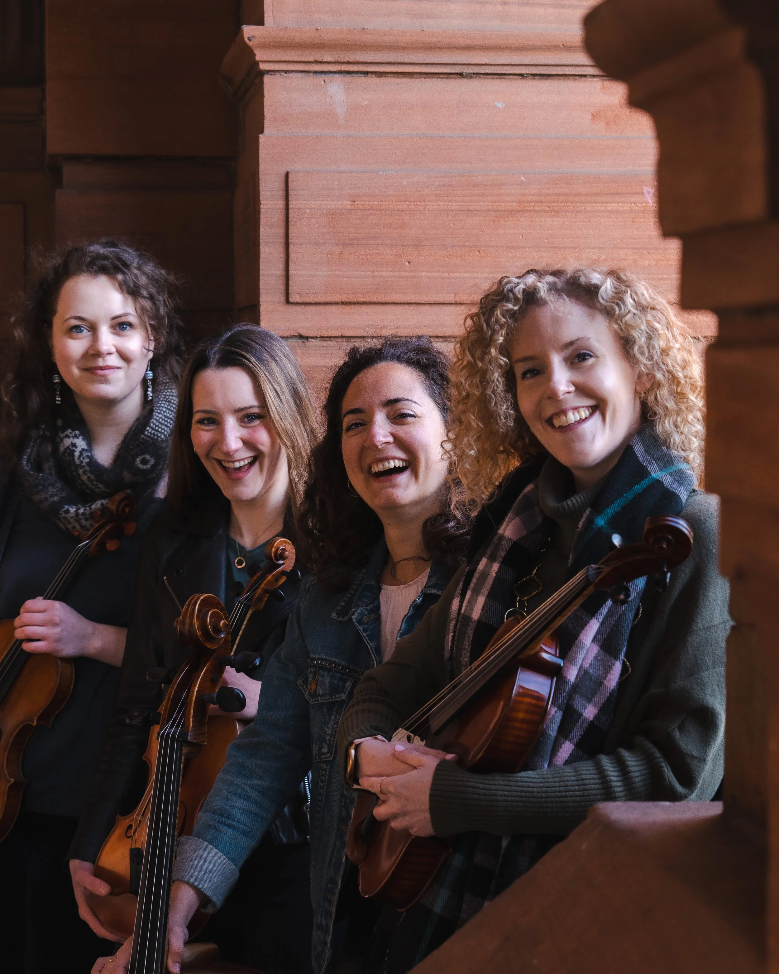 Four women standing together holding violins, smiling, in front of a stone wall.