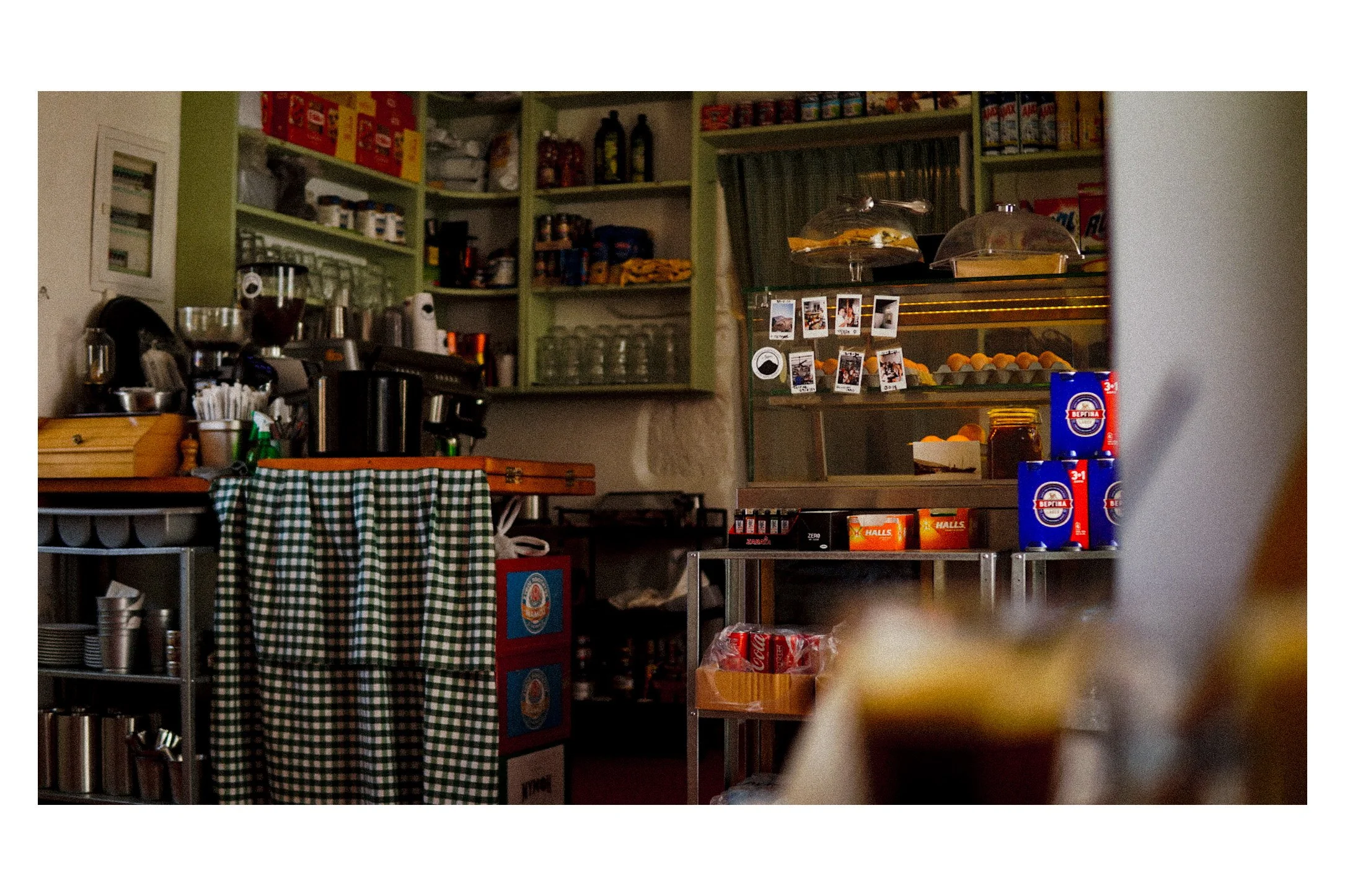 Interior of a coffee shop kitchen with shelves of jars and boxes, a glass display case with baked goods, and various coffee-making equipment.