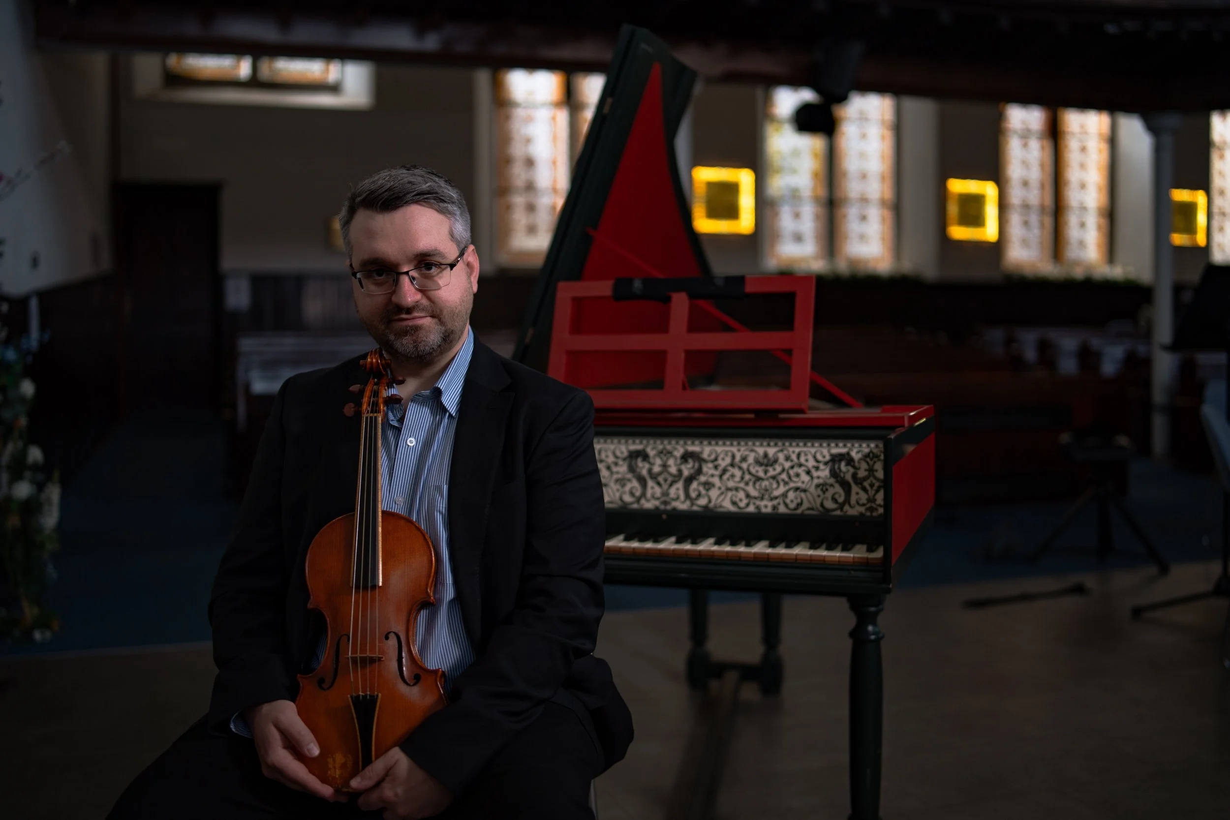 A man in a suit holding a violin sitting in front of a decorative piano in a dimly lit room with stained glass windows.