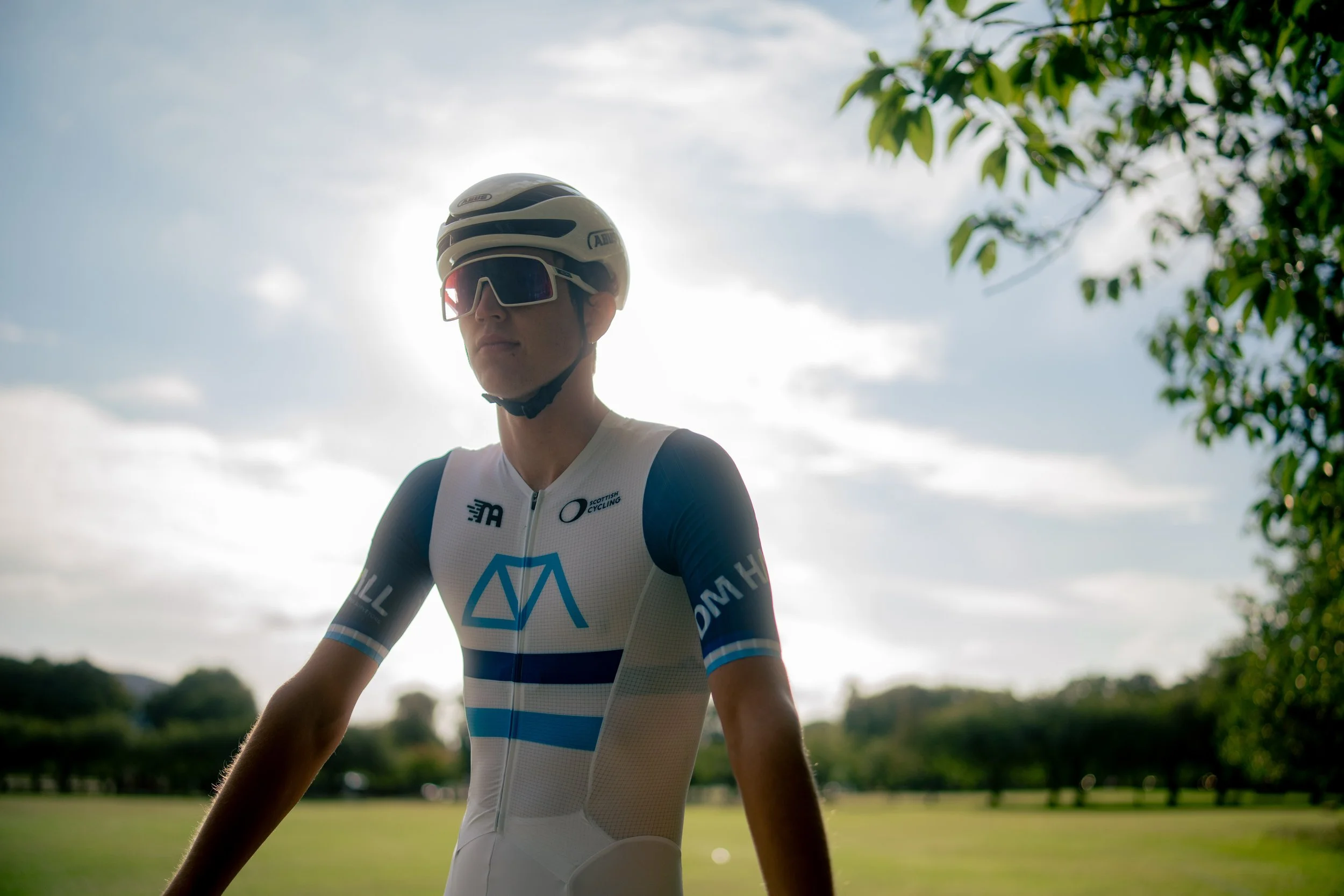 A cyclist wearing a helmet, sunglasses, and a racing suit standing outdoors in a park or open field with trees in the background, under a partly cloudy sky.