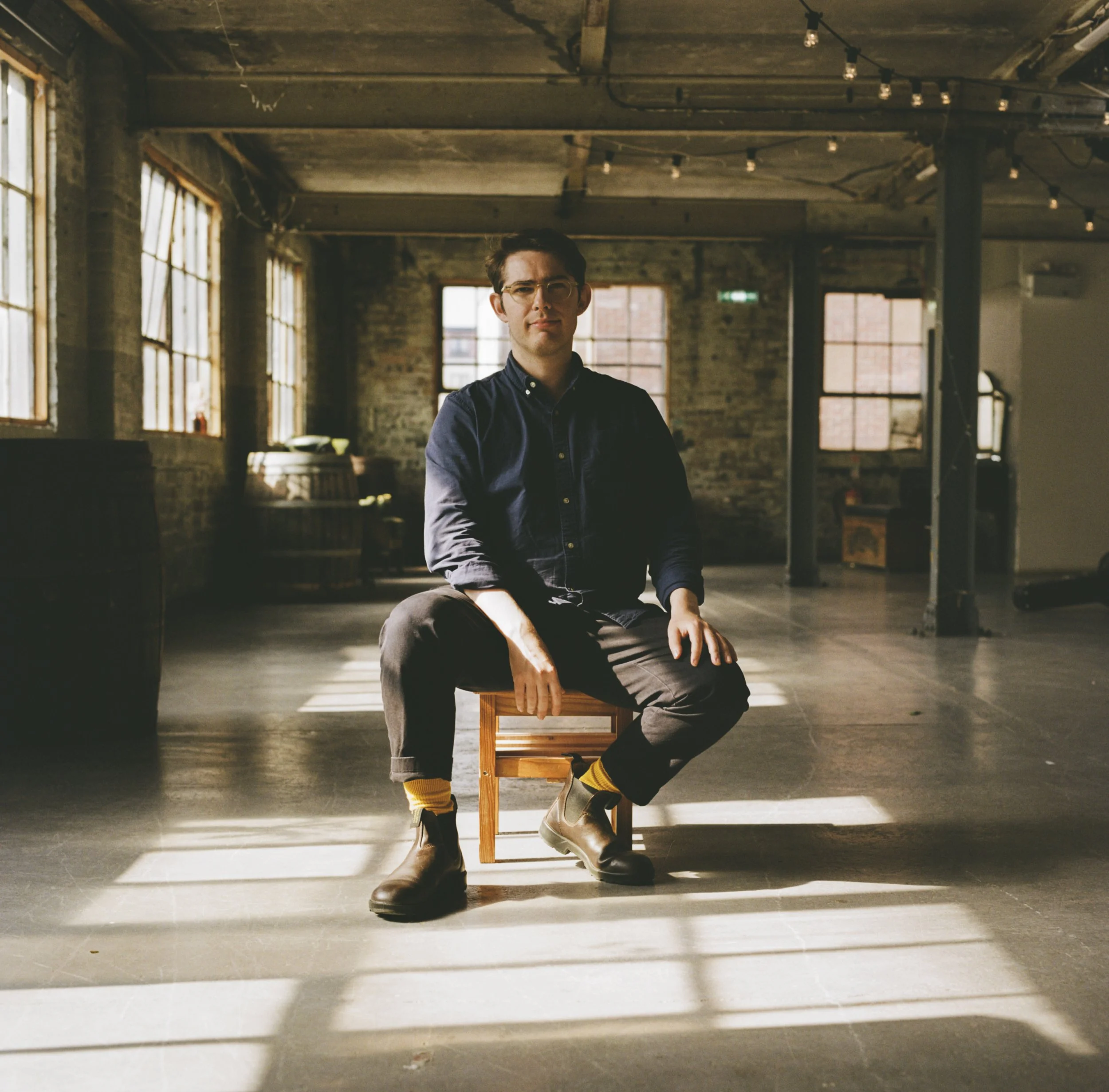 A young man sitting on a small wooden stool in a spacious industrial-style room with large windows, exposed brick walls, and string lights. He is wearing a dark shirt, black pants, yellow socks, and brown boots, with arms crossed and a serious expression.