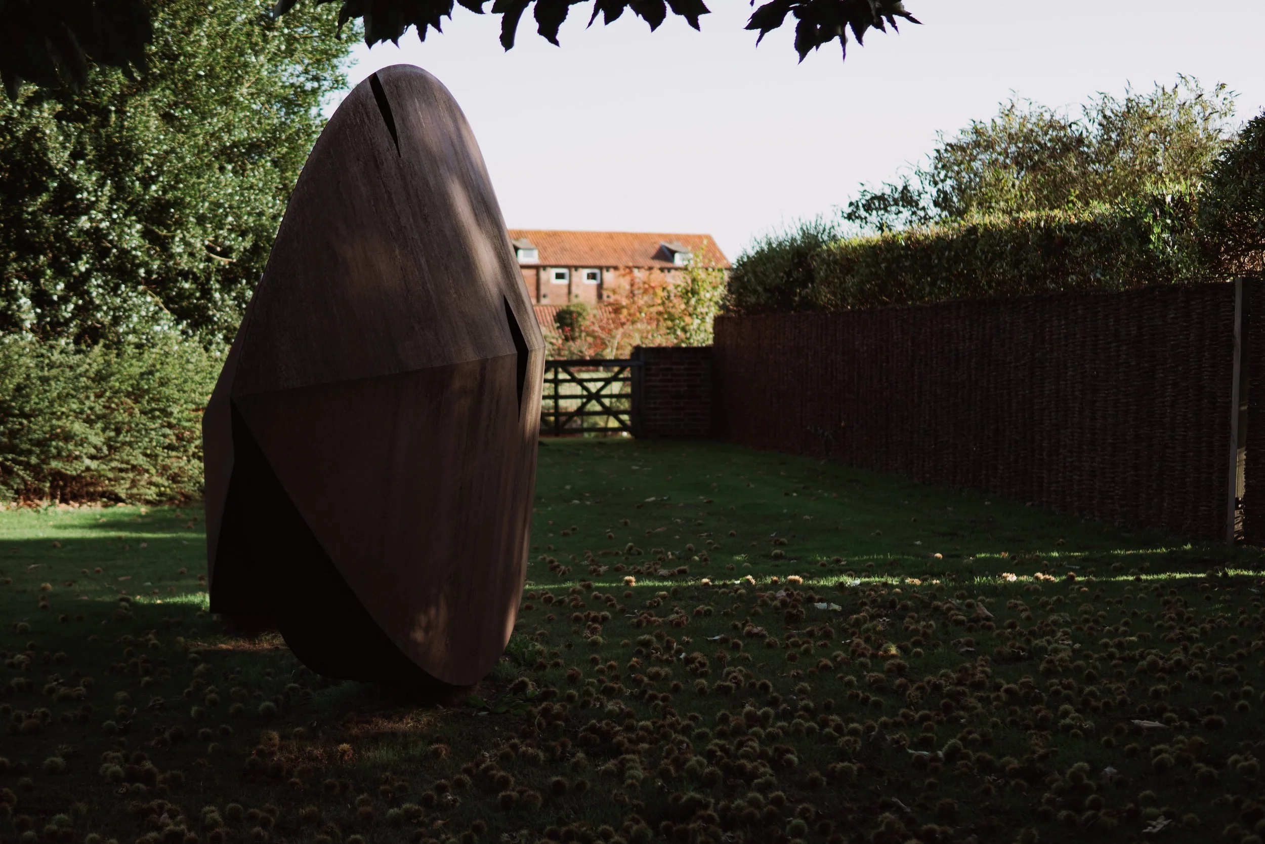 Outdoor sculpture with a geometric, polygonal shape made of dark wood or metal, situated on a grassy area surrounded by trees and a brick fence. In the background, there is a red brick house with a sloped roof.