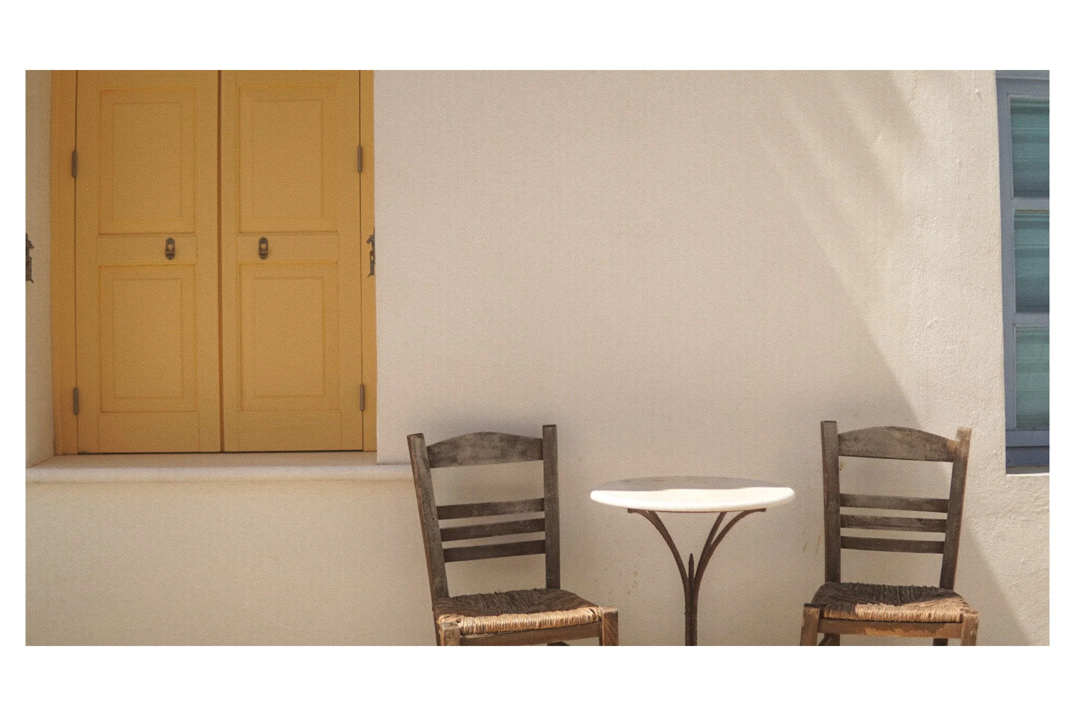 A simple outdoor scene with two wooden chairs with woven seats flanking a small round table, set against a plain white wall with a window