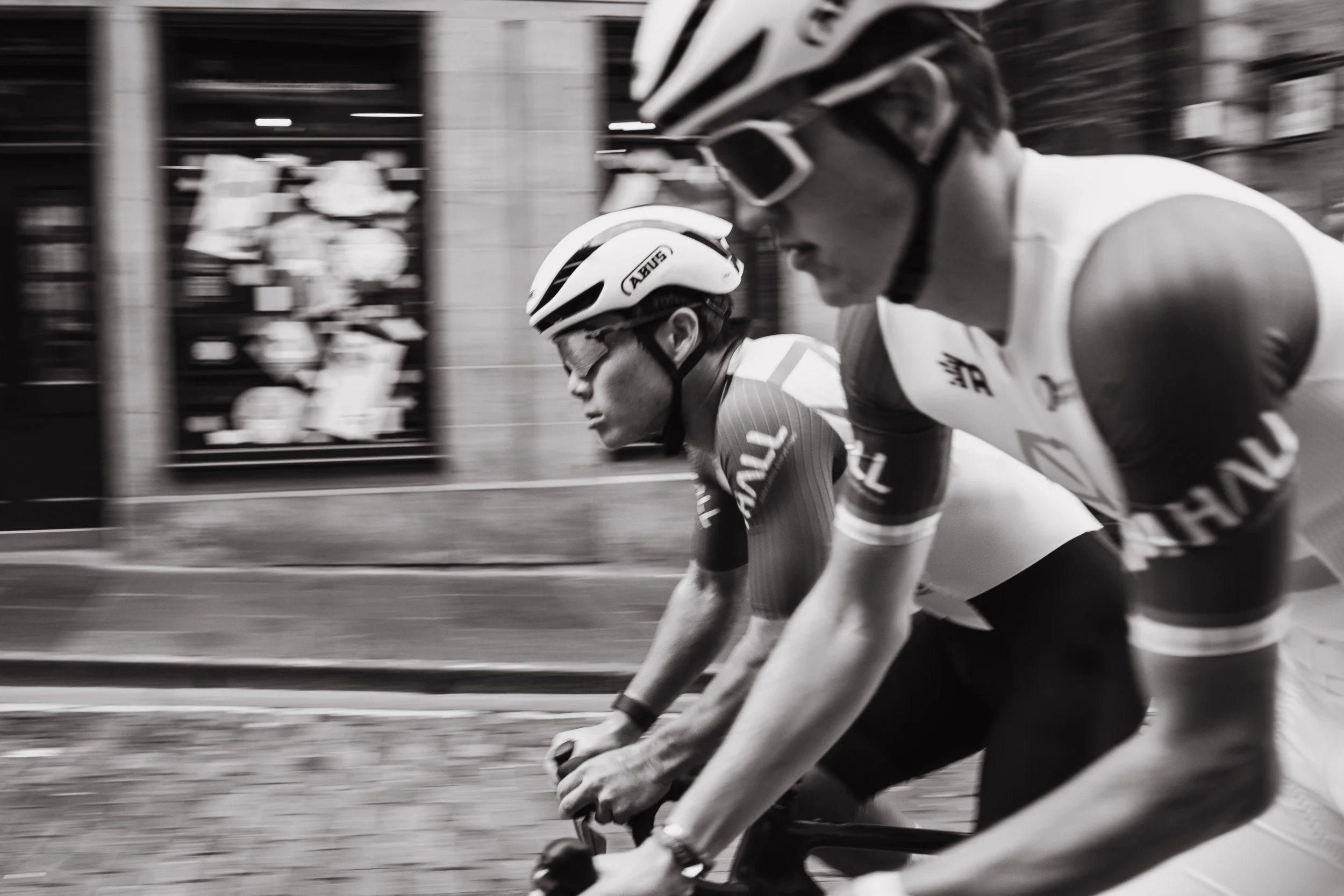 Two cyclists wearing helmets and racing gear riding side by side in a race, captured in motion on a city street.